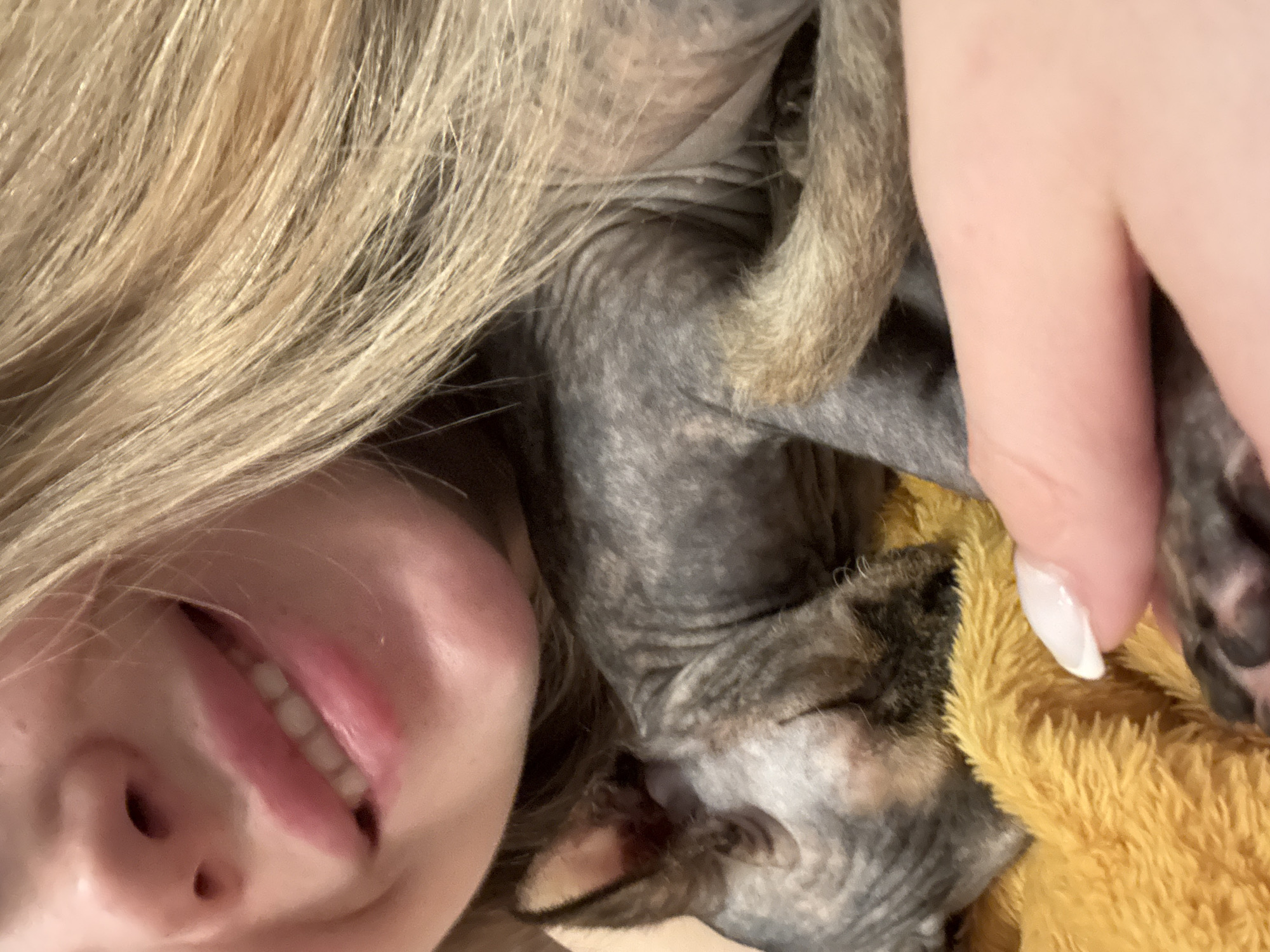 A close-up selfie depicts a person with blonde hair and pale skin, smiling with their mouth open slightly revealing teeth. Nestled against their face is a hairless dog with mottled gray and brown skin. The dog appears to be resting on or next to a yellow plush object. A fair-skinned hand with a white-painted fingernail is visible, seemingly holding or touching the dog. The location is unknown.