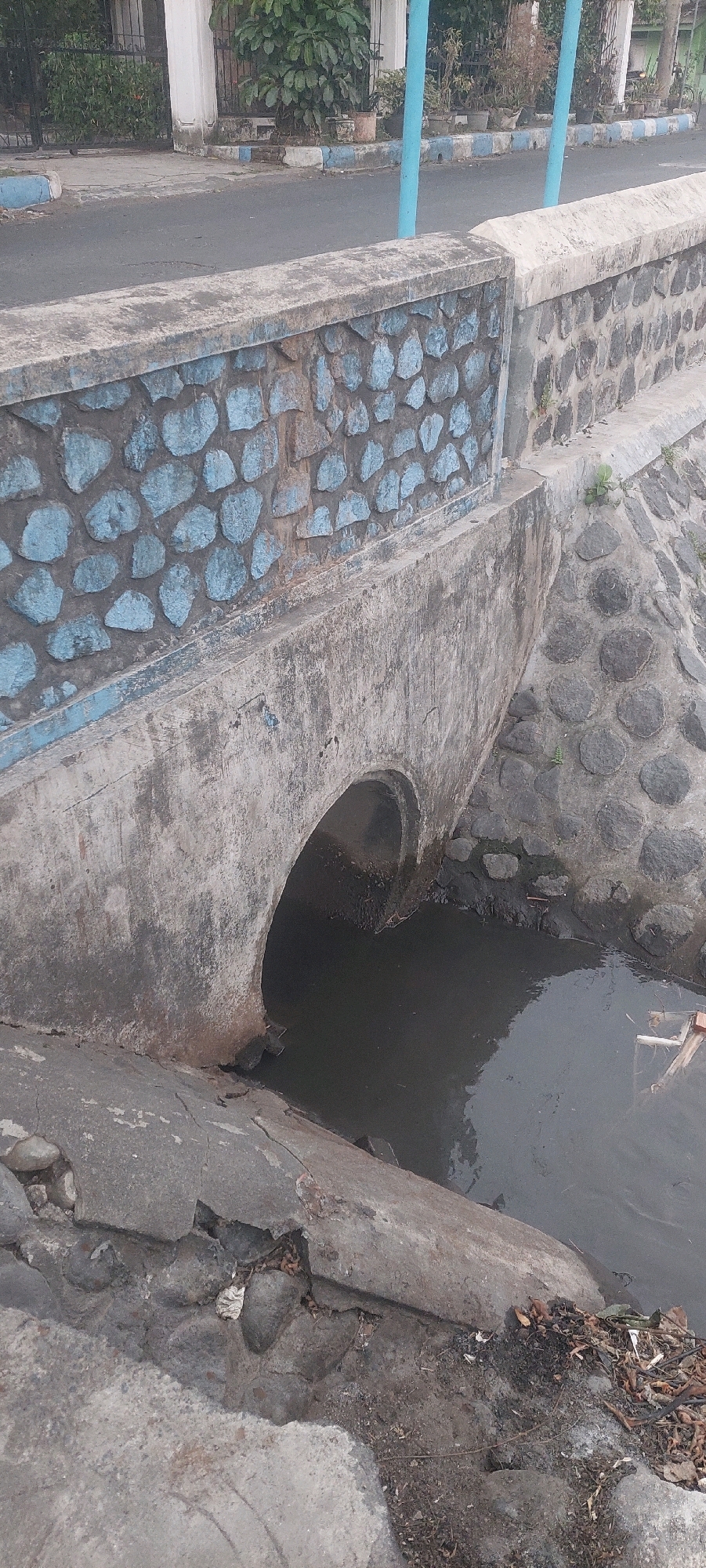 The image shows a small concrete bridge over a drainage channel in Lumajang, Indonesia. The bridge has a decorative stone facade with blue accents. The water in the channel is dark and murky, with some debris visible on the surface and along the edges. The surrounding area appears to be a street or walkway, with a road visible in the background. There are no people or animals visible in the image. The overall scene depicts a typical urban drainage infrastructure in a tropical environment.