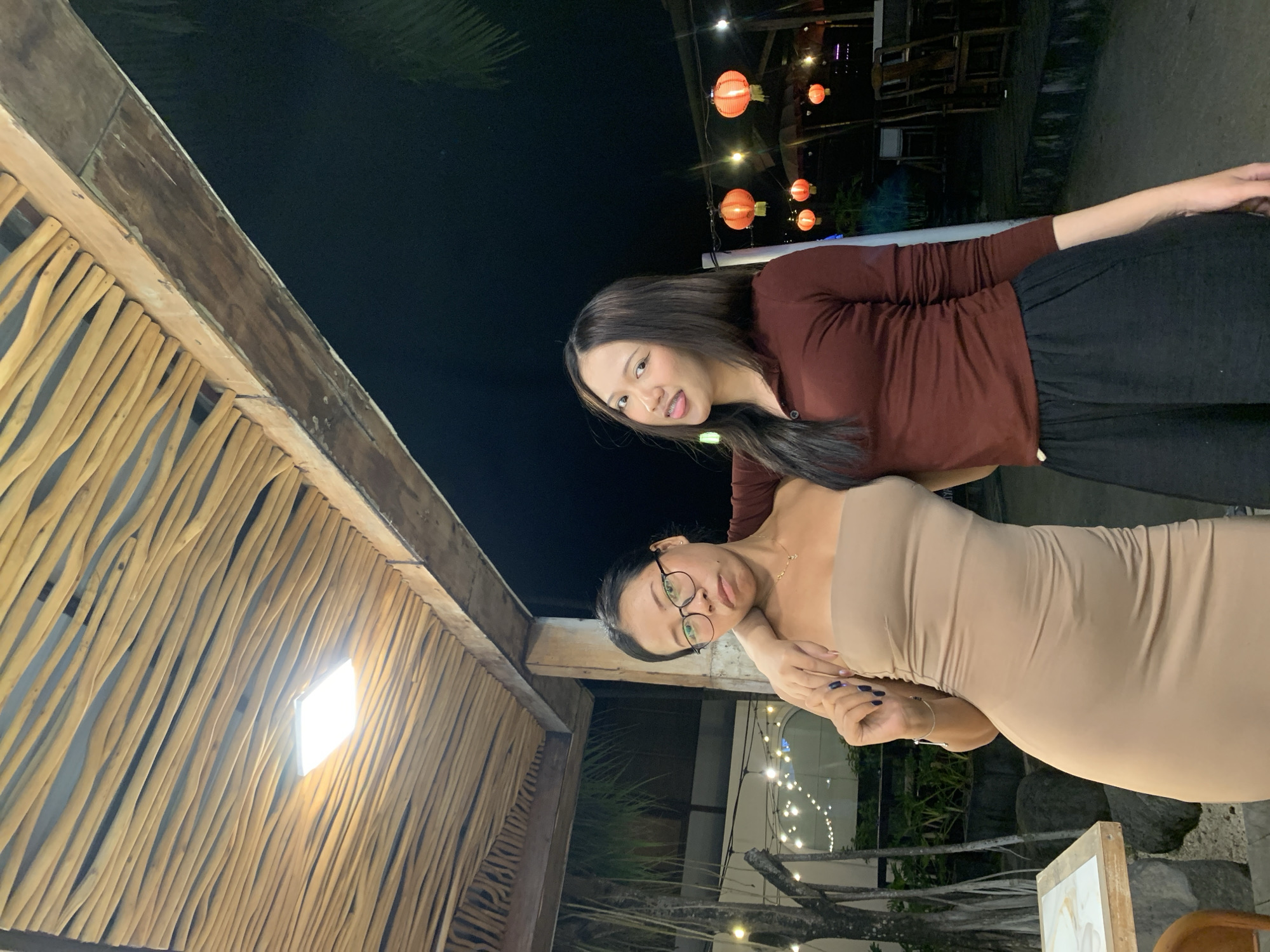 Two women are standing in an outdoor restaurant setting at night, possibly in North Kuta, Indonesia. The woman in the foreground is wearing a beige strapless dress and glasses. The other woman, in the background, is wearing a dark red top and dark pants. A wooden structure is visible to the left, with a light fixture. Lanterns and string lights are seen in the background.
