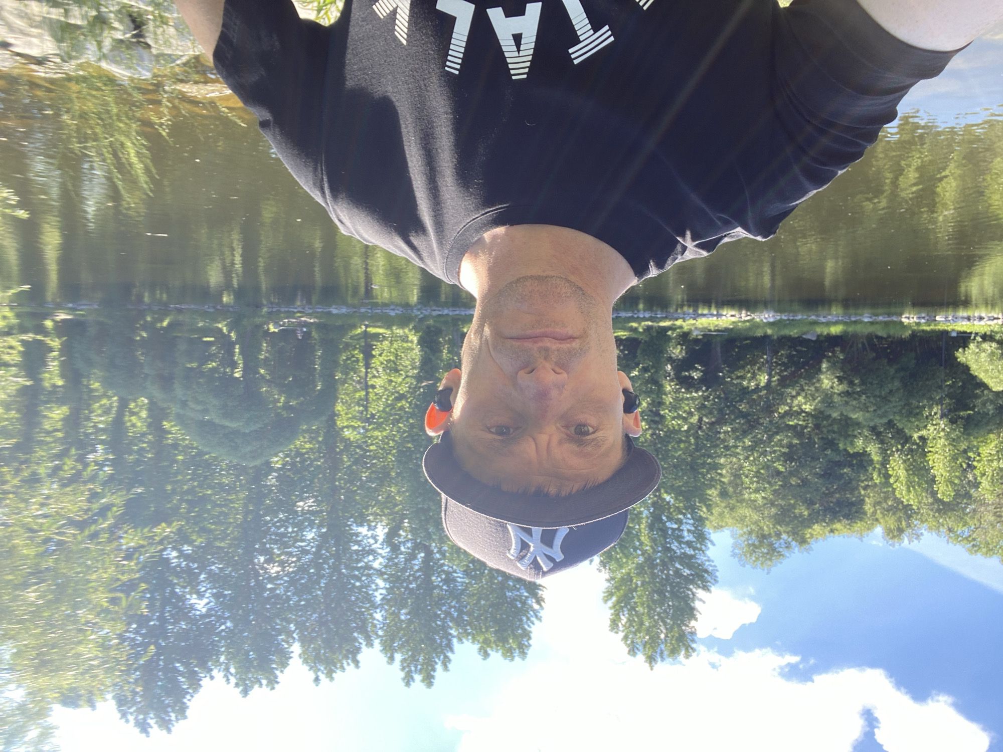 In a selfie taken outdoors, a Caucasian man wearing a black t-shirt and a New York Yankees baseball cap stands in front of a body of water. He has short, dark hair and is wearing black wireless earbuds. The man is looking directly at the camera with a neutral expression. Behind him, a calm body of water reflects the lush green trees and blue sky. The trees form a dense forest in the background, with some sunlight filtering through the leaves. The area along the water's edge is lined with rocks and grass. The overall impression is a sunny day in a park-like setting. This scene is likely in Yekaterinburg, Russia.