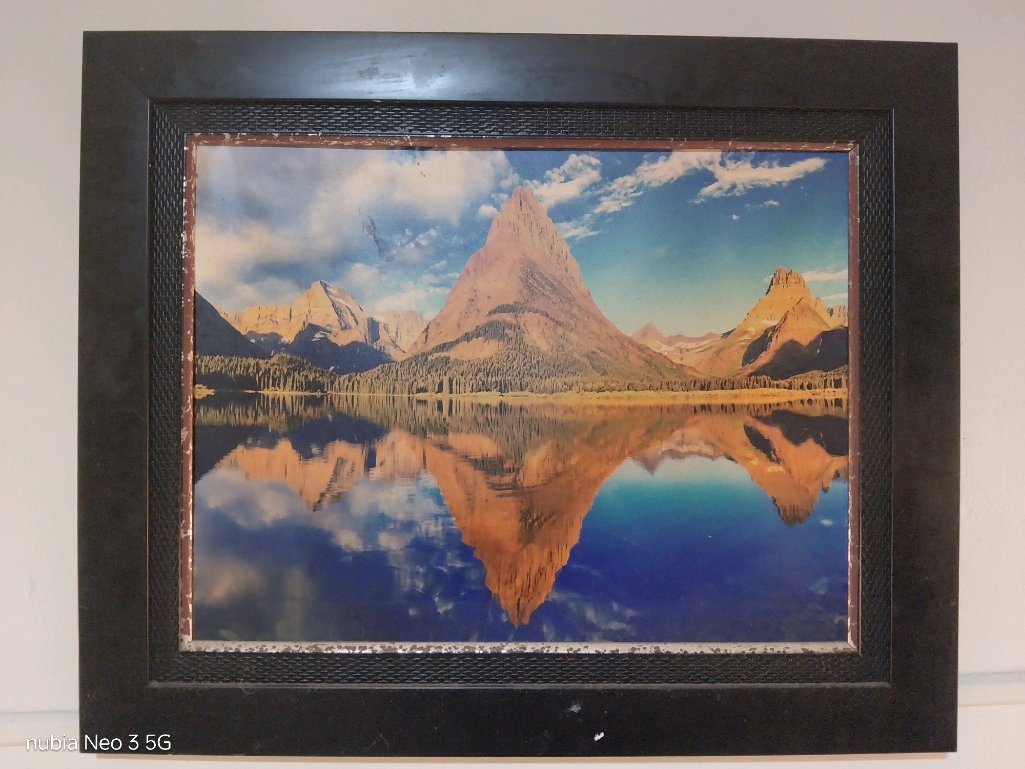 This image shows a framed picture of a mountain reflected in a lake. The frame is black with a textured pattern on the inner edge. The picture itself depicts a large, rocky mountain with snow-capped peaks, set against a blue sky with fluffy white clouds. A dense forest lines the shore of the lake, and the mountain's reflection is clearly visible in the calm water. The photograph appears to be taken indoors, likely in Tembilahan, Indonesia, though the specific location within the city is not discernible from the image. The scene is static and does not include any people or animals.