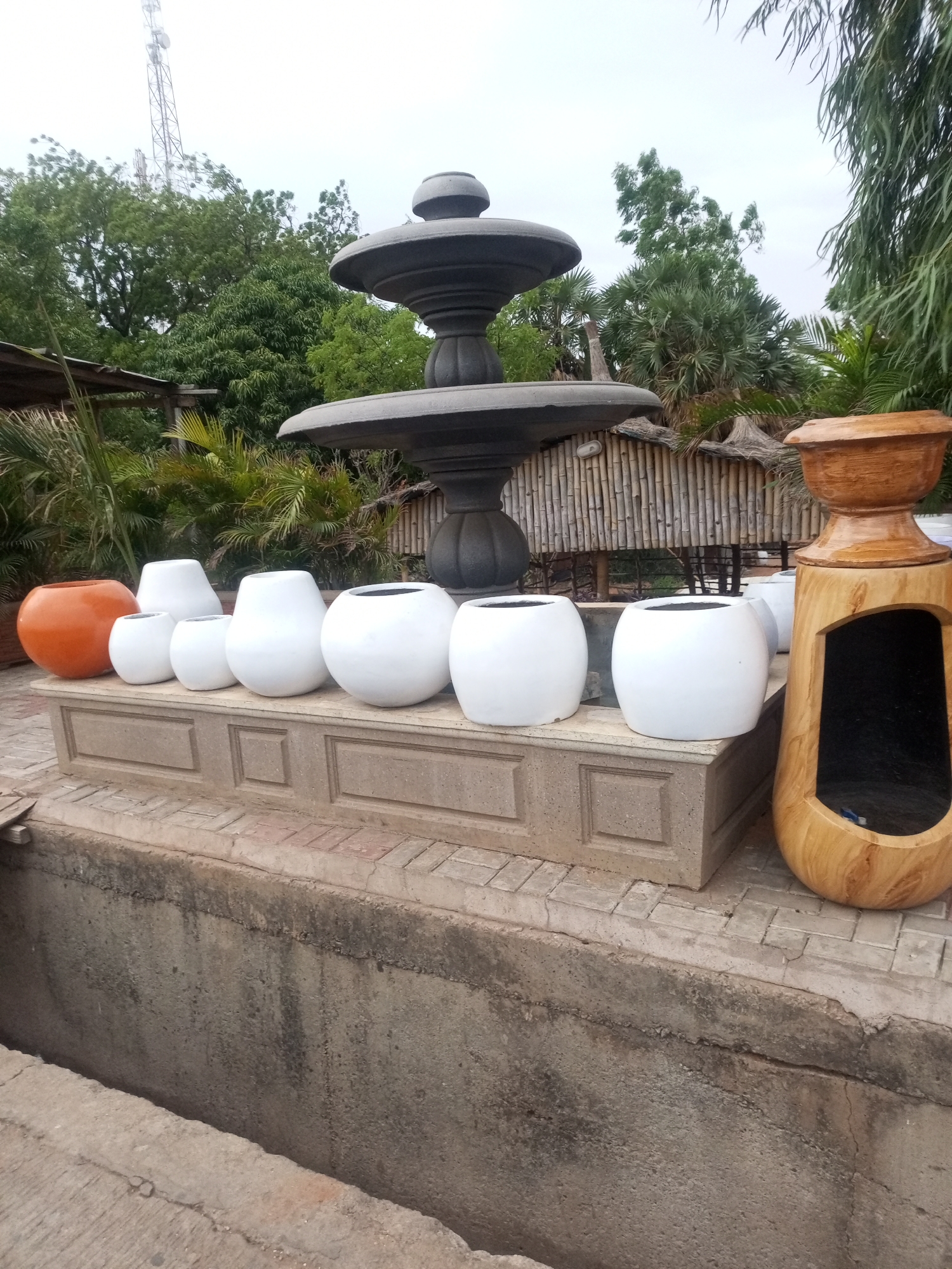 The image captures a scene with several decorative objects arranged outdoors in an unknown city, unknown. A collection of round, white and orange planters of varying sizes sit atop a raised concrete platform. Adjacent to them is a tall, light-colored wood structure featuring a recessed oval opening. Behind these objects stands a multi-tiered water fountain of a darker tone. In the background, there is a bamboo structure and various trees. A tall communication tower is visible above the trees. A concrete wall with a textured surface runs along the foreground, partially obscuring the view.