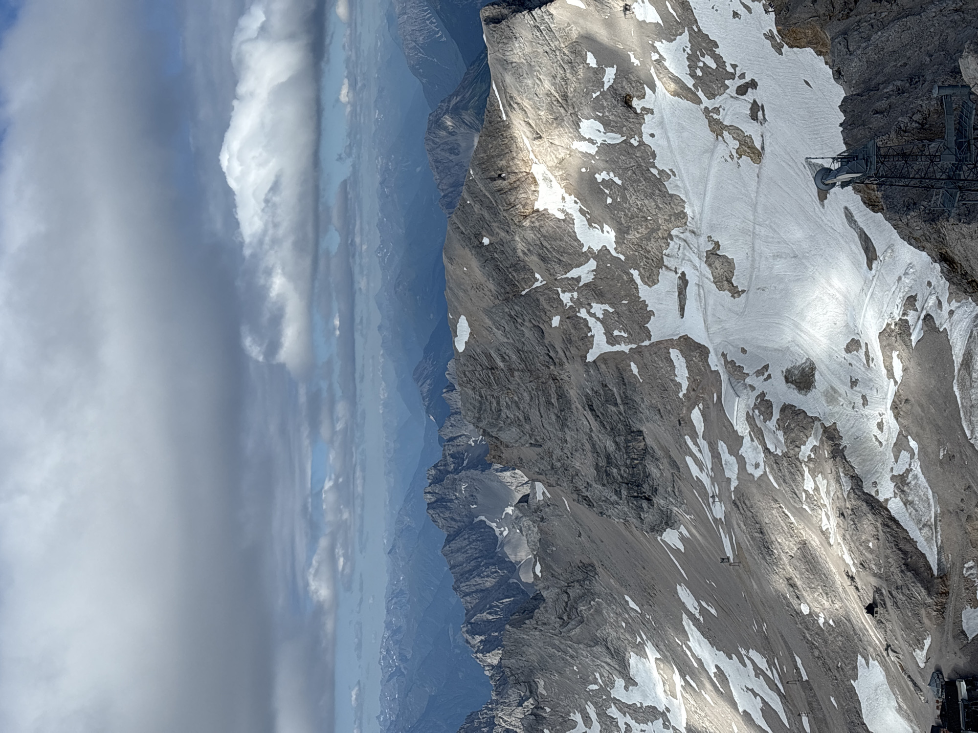 This is a high-angle, full shot of a rugged, snow-dusted mountain range under a cloudy sky. The mountains are predominantly grey rock with patches of white snow, suggesting a high altitude. In the foreground, a metal tower with various antennas and dishes is visible on a rocky peak. The terrain slopes downwards, revealing more snow and rock formations, with some ski tracks visible on the snow. A small, dark building sits on the mountainside further down. In the far distance, layers of receding mountain ranges fade into a hazy blue. The sky is filled with dramatic, grey and white clouds, with patches of blue sky visible in between. The lighting suggests it is daytime, with some areas of the mountains in shadow and others illuminated by sunlight. While the image depicts a mountainous landscape, there is no specific indication of the city of Ehrwald, Austria, within the frame.