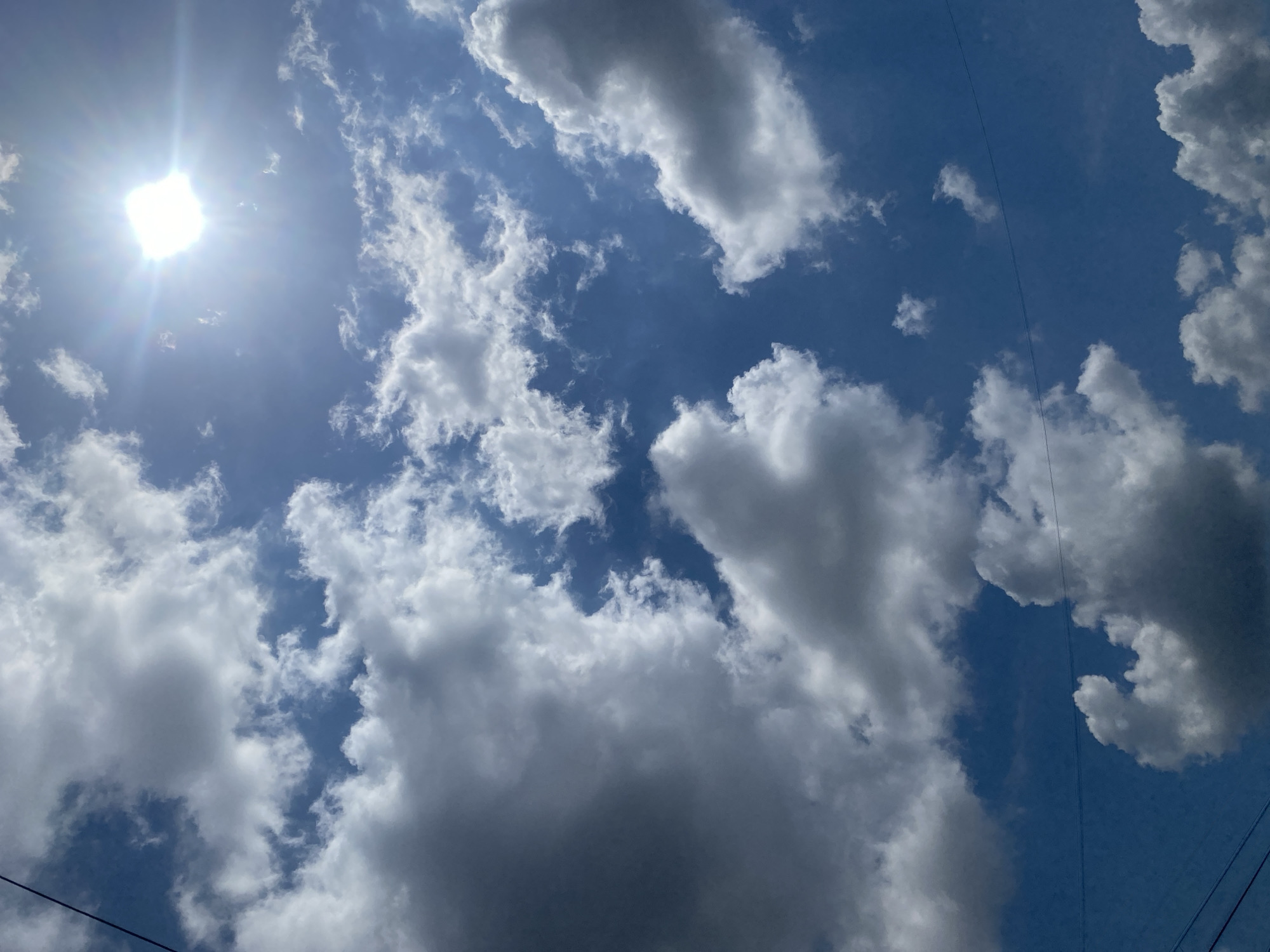 A clear blue sky is visible with scattered white and gray clouds. The sun is brightly shining from the upper right corner, casting a lens flare. In the foreground, the tops of power lines are visible. This scene is located in Berangas Timur, Indonesia.