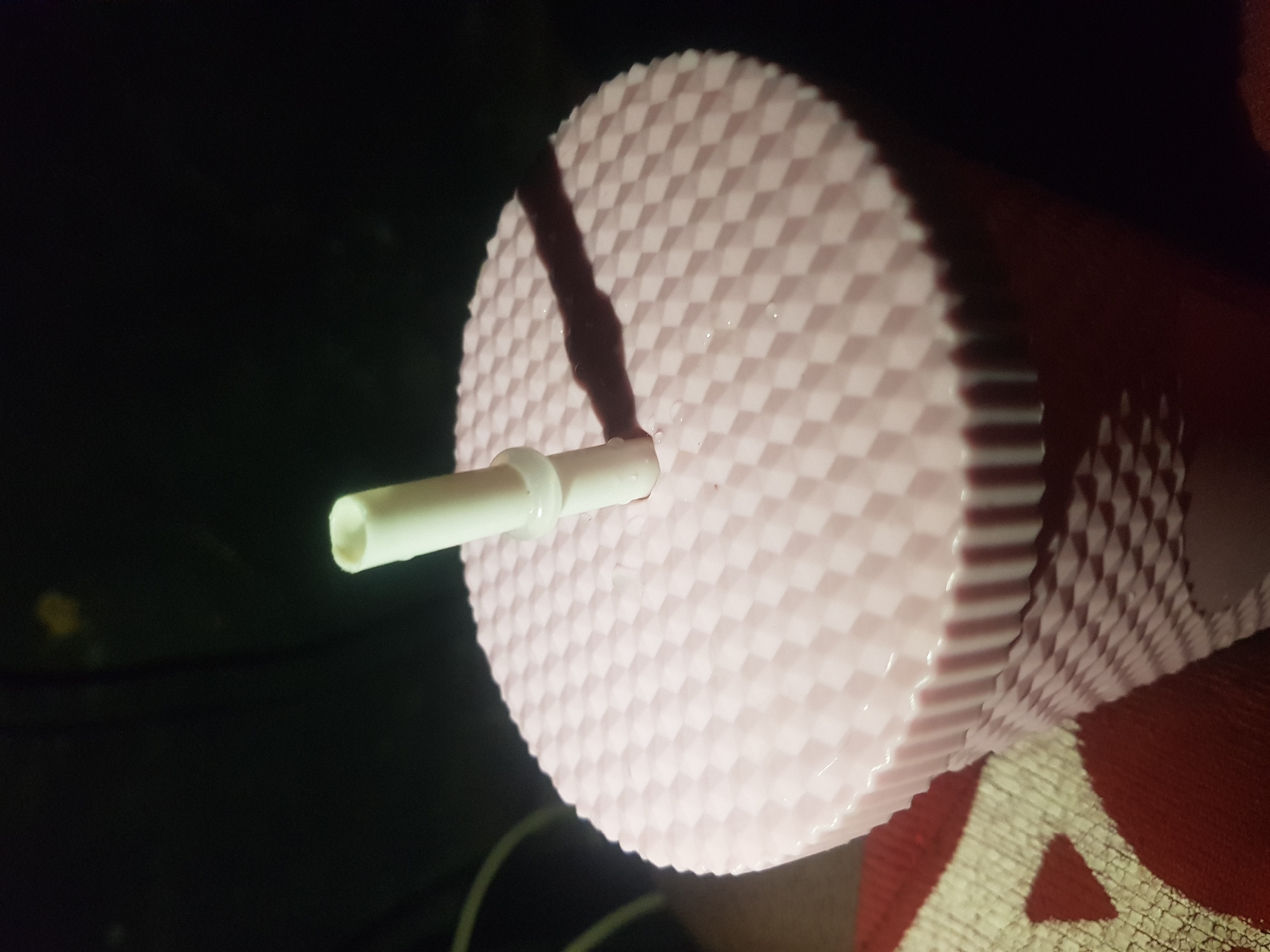 The image shows a close-up of the top of a pink, diamond-patterned tumbler with a white straw inserted through the lid. The straw has a small, clear plastic ring around it. The background is dark and out of focus. A shadow of the straw is cast across the textured surface of the tumbler lid. The scene appears to be indoors, possibly in a car or a dimly lit room, given the lighting conditions and the visible texture of what might be a seat or fabric in the lower left of the frame. There are no people visible in the image.