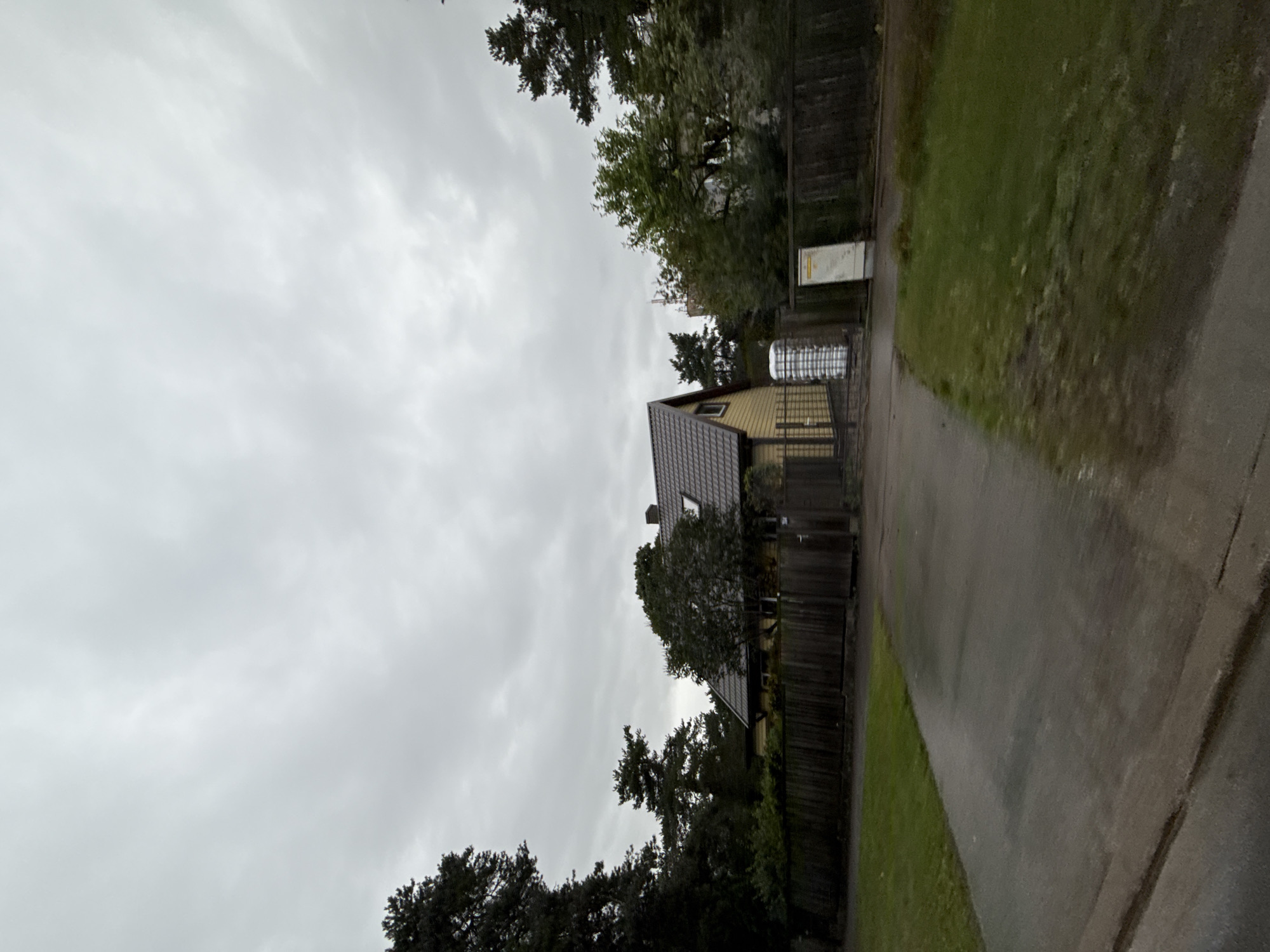 The image depicts a residential scene under a heavily overcast, gray sky. In the center, a yellow wooden house with a dark tiled roof is visible. The house has a small dormer window on the roof. A dark wooden fence runs across the front of the property, with a gate to the right of the house. Next to the gate is a white cylindrical object, possibly a water tank or storage container. To the right of this, a light-colored rectangular utility box with some text on it is affixed to the fence. In front of the house and fence, there is a paved driveway or road, and to the left of that, a strip of green grass. Tall pine trees and other deciduous trees are present on the property and surrounding the house, some of which are dark and imposing. In the distance, through the trees, a tall structure with antennas is visible, suggesting it could be a telecommunication tower. The overall impression is that of a suburban or semi-rural residential area in Tallinn, Estonia, likely during a cool or damp day. There are no people visible in the image.