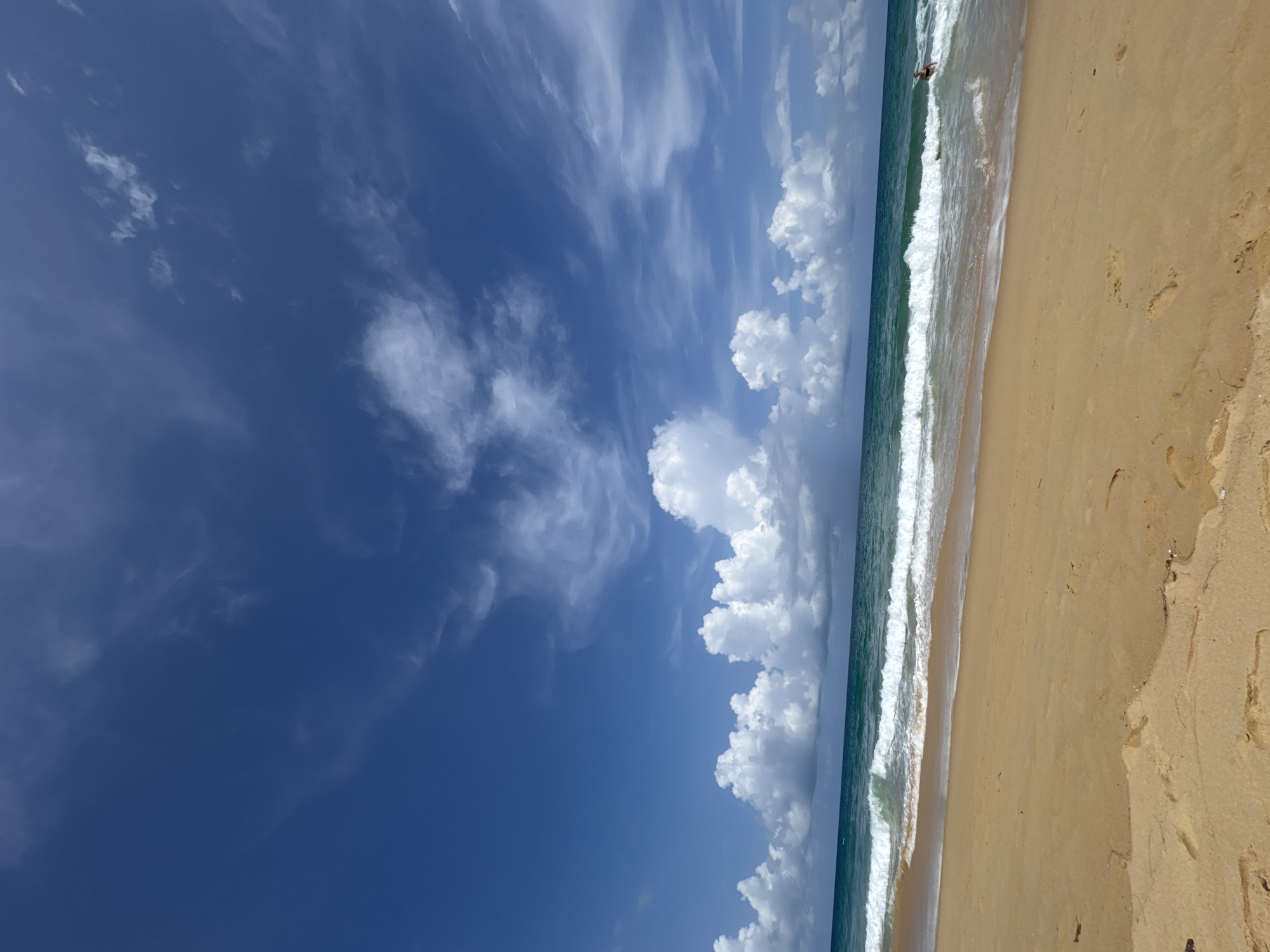 This image depicts a wide, sandy beach in Karon, Thailand, under a bright blue sky filled with white, fluffy clouds. The ocean is a vibrant turquoise, with moderate waves rolling onto the shore. In the distance, a single person can be seen swimming or playing in the water. The foreground is dominated by the golden sand, with footprints visible, suggesting recent activity on the beach. The overall scene conveys a sense of a peaceful and beautiful day at the beach.