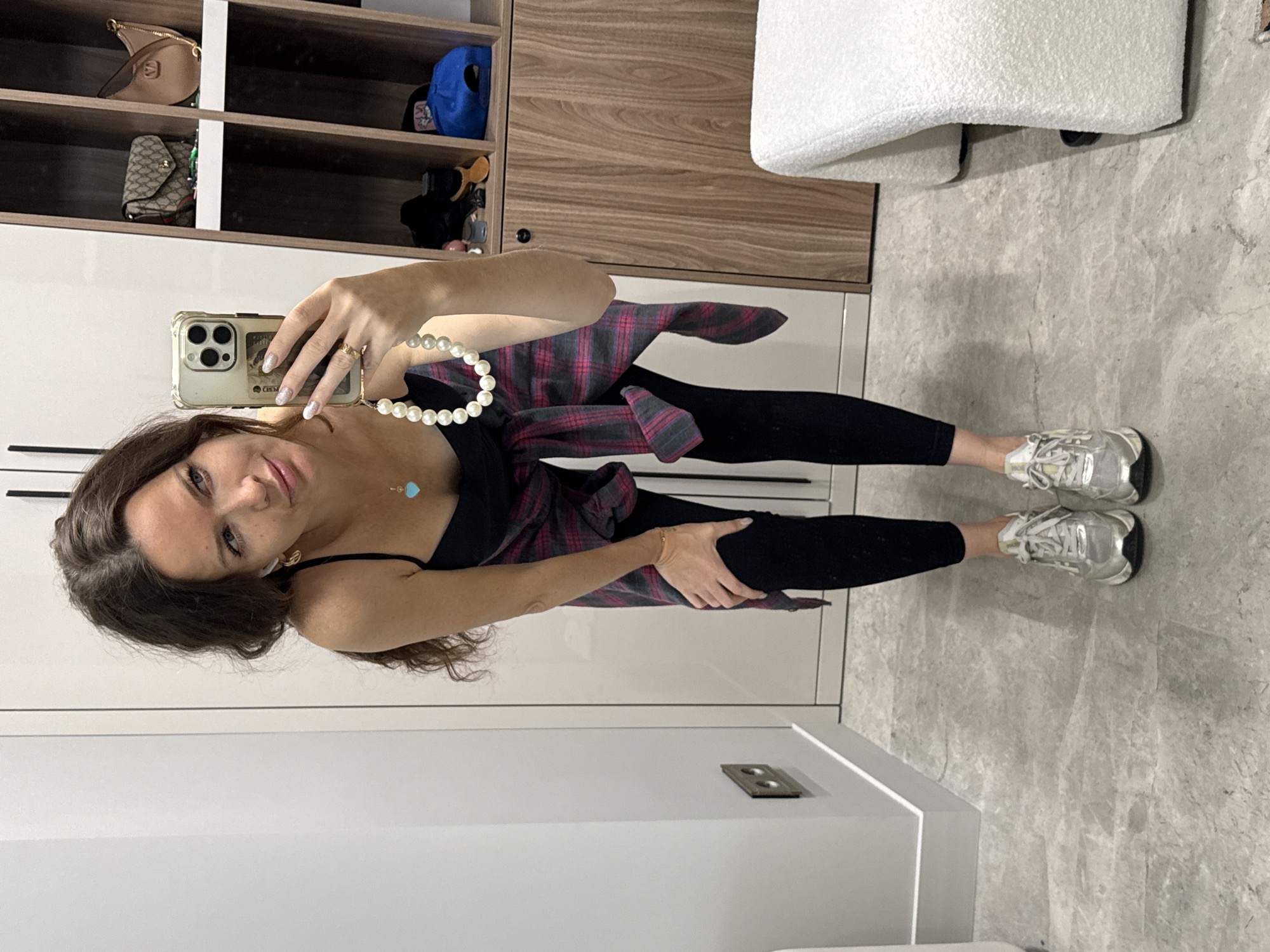 A woman with long, dark, wavy hair is taking a mirror selfie. She is wearing a black tank top, black leggings, and silver sneakers. A red and gray plaid shirt is tied around her waist. She has a pearl necklace with a blue heart pendant and a gold ring on her right hand. The background shows a white wall, a wooden shelf unit with bags and a blue baseball cap, and a white armchair. The floor is tiled with a gray marble pattern. The location appears to be a modern dressing room or closet area.