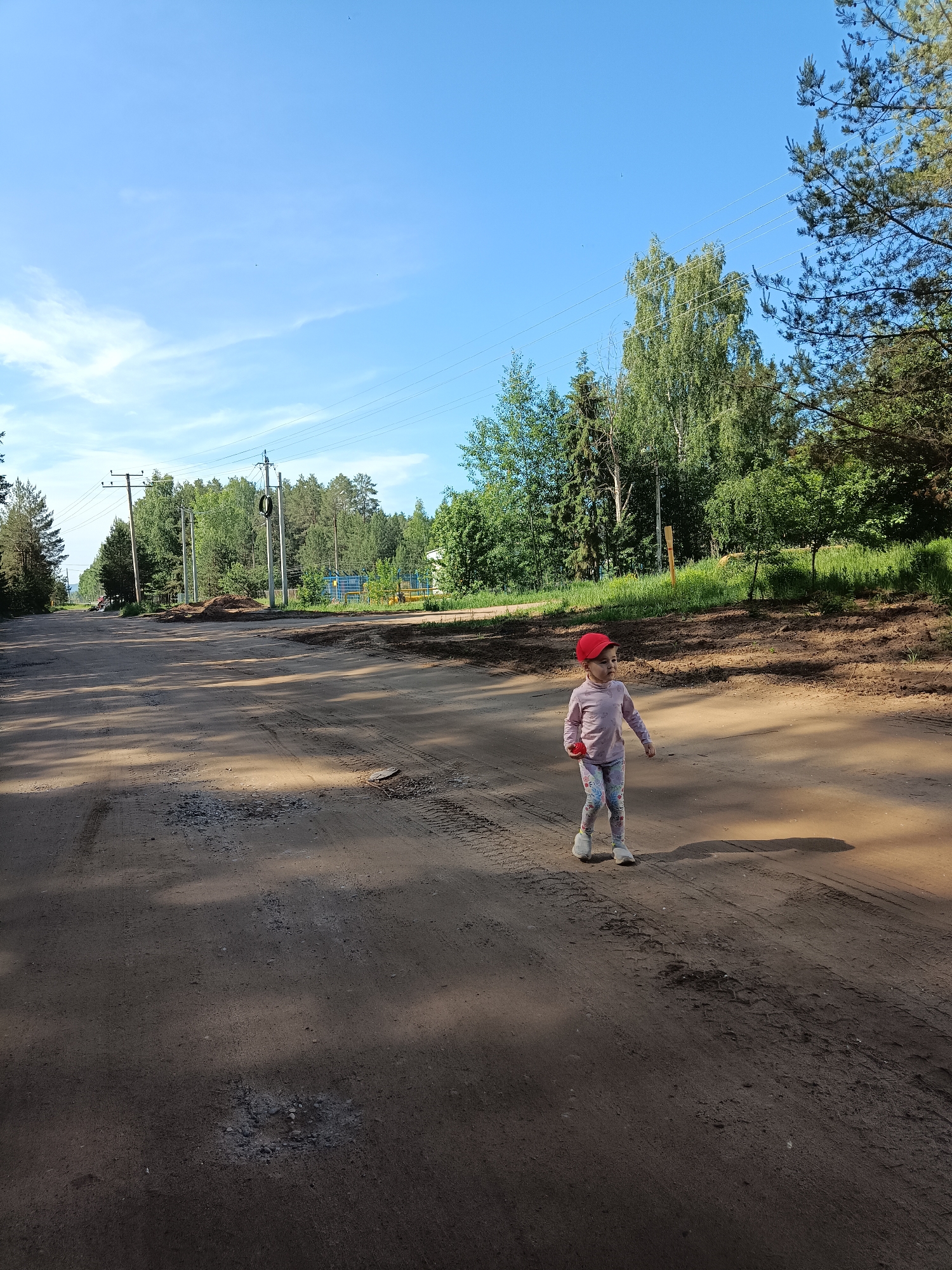 A young child, appearing to be a girl, is captured walking or standing on a dirt road in a sunny, semi-rural setting, identified as Noviy, Russia. The child, seen from the waist up in the mid-right of the frame, wears a red baseball cap, a light pink long-sleeved top, patterned leggings, and white sneakers, holding a small red object in her left hand. Her expression is neutral as she looks towards the left side of the frame.
The unpaved road, which dominates the lower half of the image, is covered in dirt, gravel, and shows numerous tire tracks and some potholes. Long shadows fall across the road from the right, suggesting it is either morning or late afternoon. To the left, the road extends into the distance, lined by power poles carrying multiple overhead lines.
The background features abundant green foliage, including tall pine trees and leafy deciduous trees, creating a dense forest line on both sides of the road. A clear, bright blue sky with a few faint wispy clouds is visible above the tree line. In the mid-ground, on the left side of the road, piles of earth or construction materials are present. Faintly visible through the trees on the right is a blue structure. The scene is bright and well-lit, conveying a calm outdoor atmosphere. There is no visible text in the image.