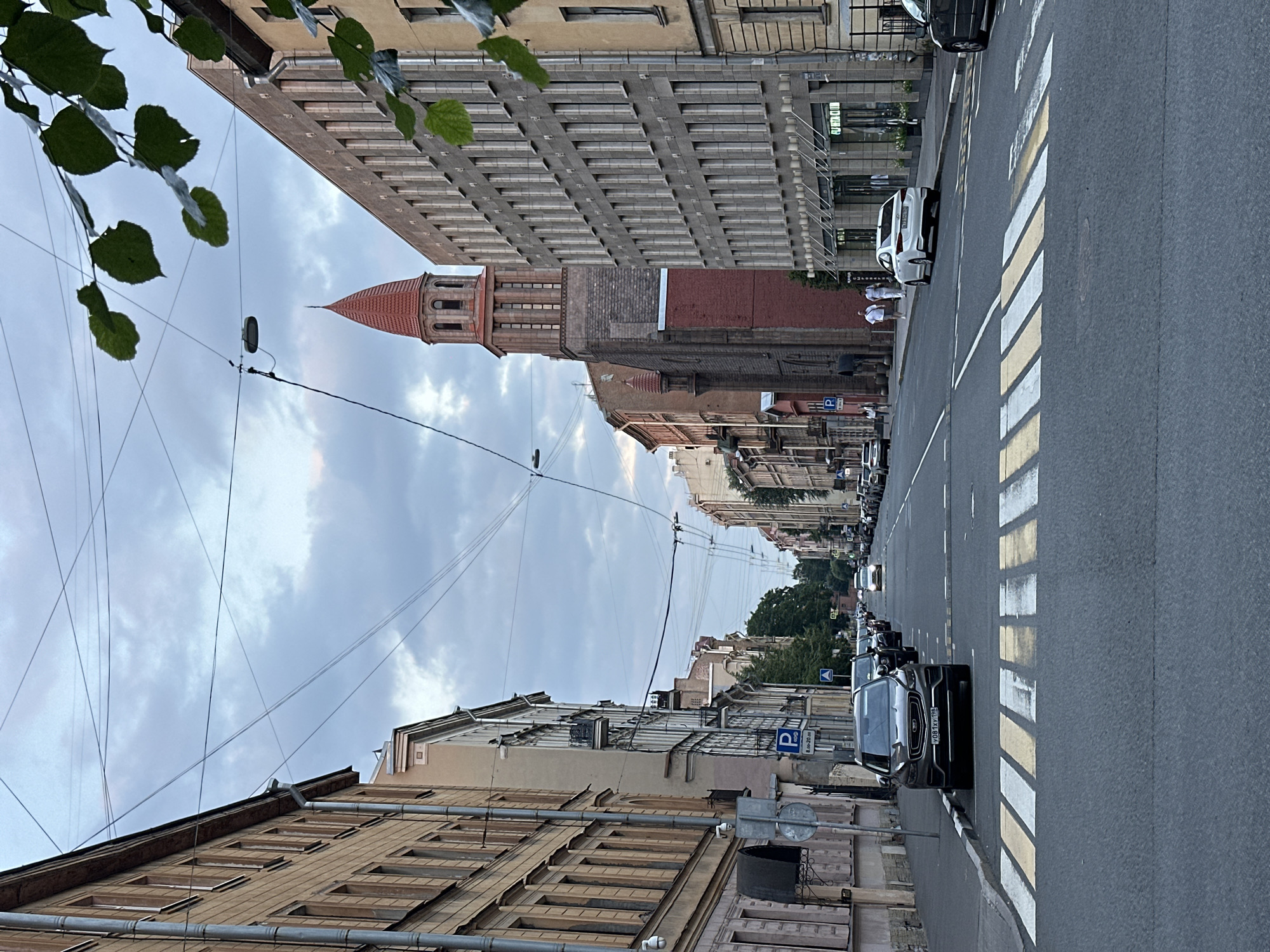 The image depicts a street scene in Saint Petersburg, Russia. The view is looking down a straight, paved street lined with multi-story buildings on both sides. A pedestrian crosswalk with yellow and white stripes is prominent in the foreground. Several cars are parked along the right side of the street, and others are driving on the road. On the left side, there is a parking sign. In the distance, a distinctive church tower with a red conical roof rises above the buildings. The sky is overcast with clouds, and some tree leaves are visible in the upper right corner. Two individuals are walking on the sidewalk in the middle distance on the right side of the street. The architecture suggests an urban environment typical of Saint Petersburg.