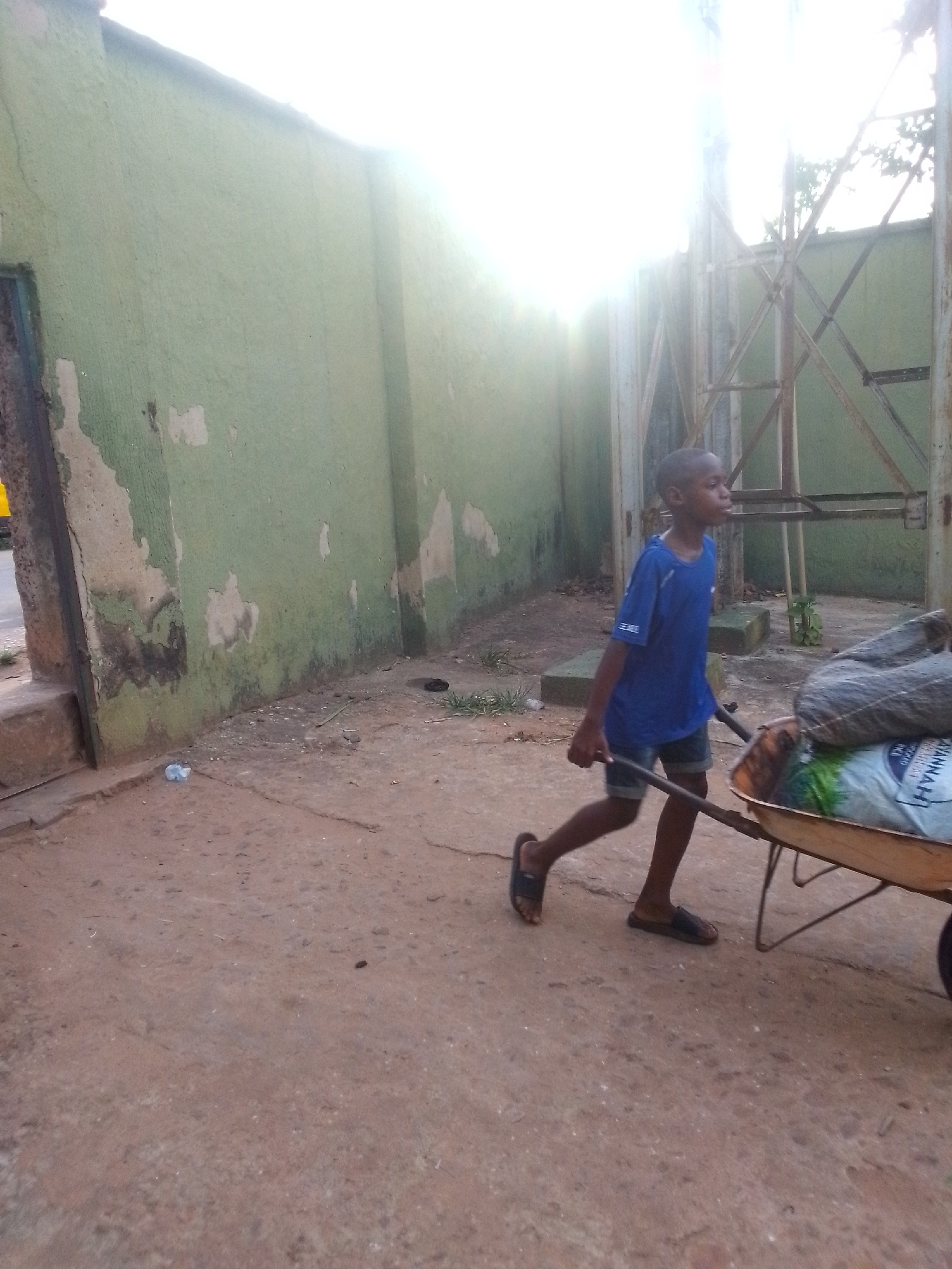 A young boy wearing a blue t-shirt and dark shorts, and sandals is pulling a wheelbarrow filled with burlap sacks along a dirt and concrete area. He is walking towards the right side of the frame. To the left is a peeling, pale green wall with a dark doorway. Behind the boy is a tall metal structure with intersecting diagonal supports. The background is bright, with a haze obscuring details. The location appears to be a street in Onitsha, Nigeria.