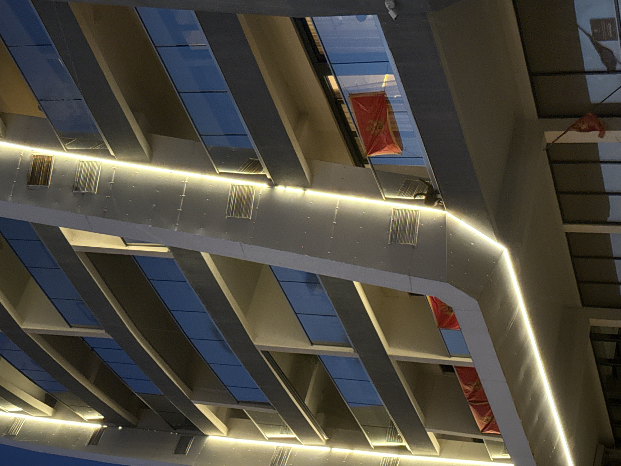 The image shows a view from below of a modern architectural structure, likely a building or overpass, with a series of repeating geometric patterns. The structure is illuminated with linear LED lights that accentuate the architectural design. Within the openings are visible the flag of Montenegro. The location is Bar, Montenegro. The image is taken from a low angle, looking upwards. No people are visible in the image.