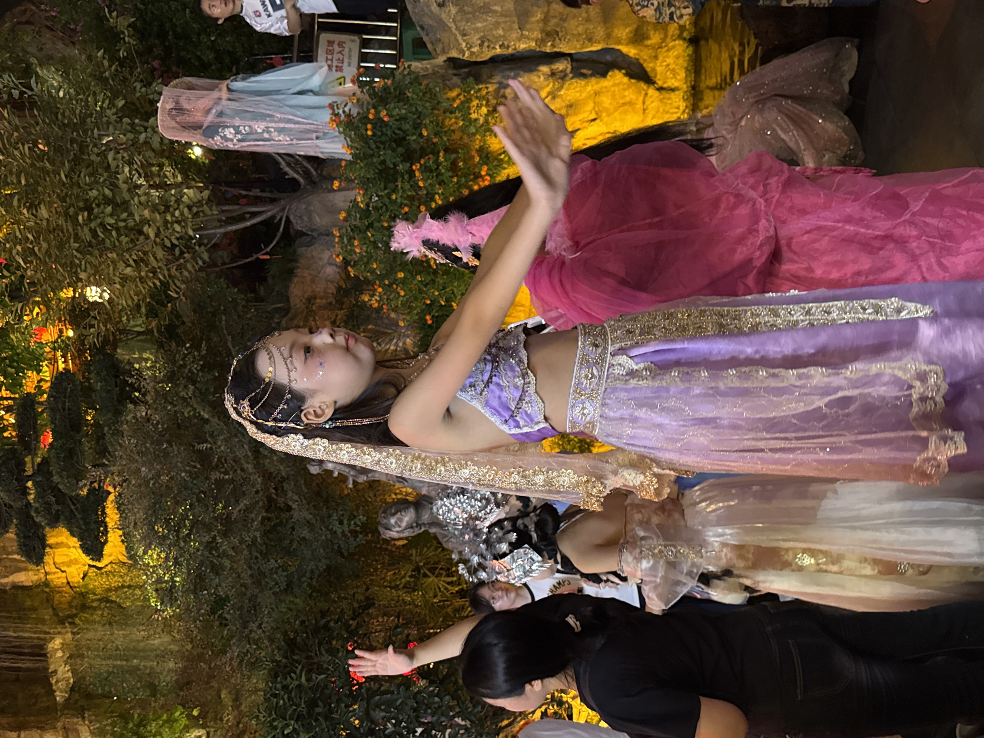In Yuzhong District, China, a young girl in a purple and gold costume, possibly performing, is in the foreground with her right arm extended. She has a jeweled headpiece and makeup on her face. To her left, a woman in a black shirt and jeans looks up with her left arm raised. Behind the main performer, another person wears a pink voluminous dress. In the background, a person in a light blue traditional outfit with a veil stands partially obscured by foliage, and further right, a person in a white t-shirt observes the scene. The environment appears to be an outdoor garden or performance space with artificial rock formations, lush greenery, and illuminated water features.