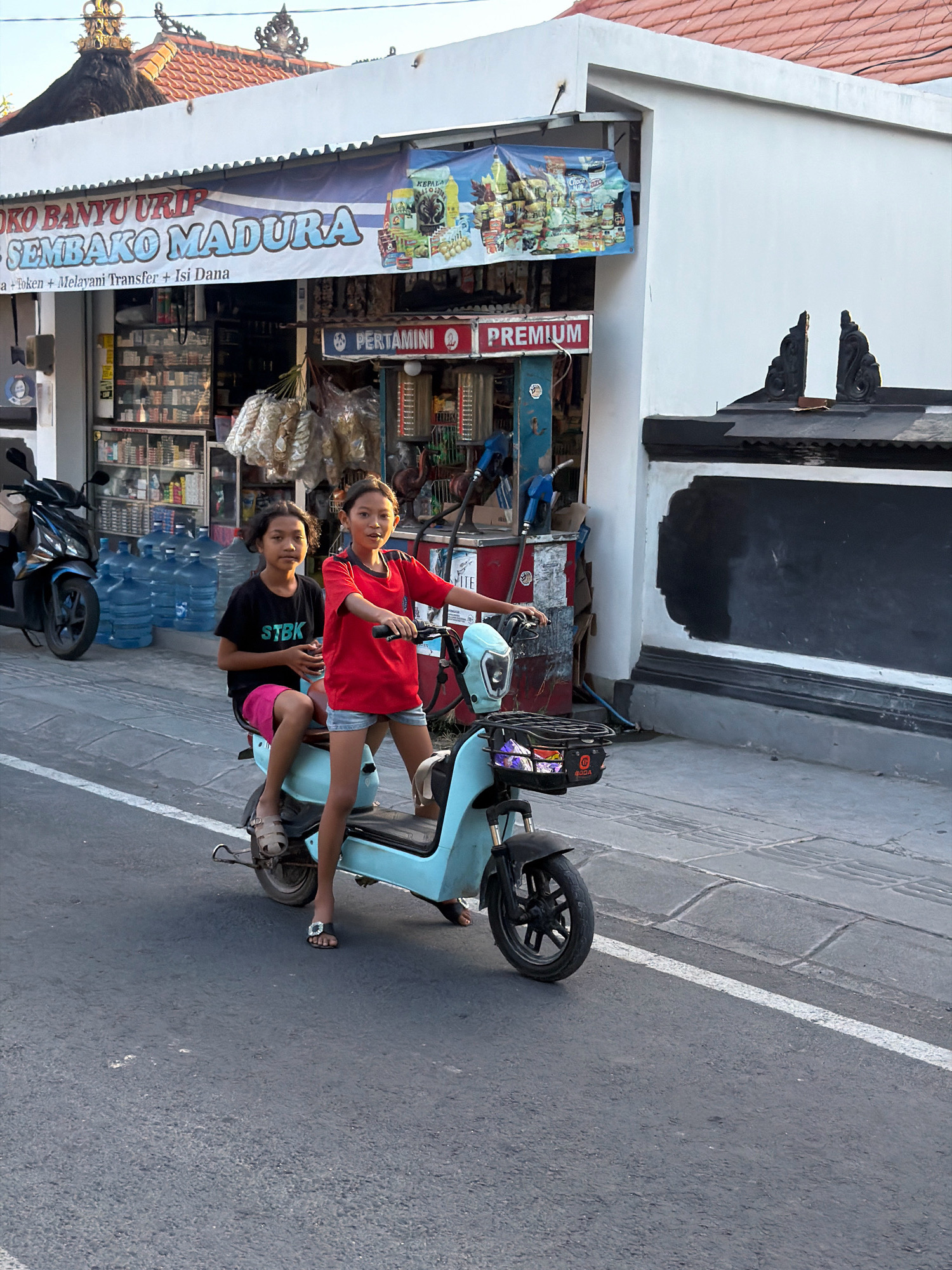 Two young girls are riding on a light blue electric scooter on an asphalt road in front of a storefront. The girl in the front is wearing a red t-shirt and denim shorts, and the girl in the back is wearing a black t-shirt with "STBK" printed on it and pink shorts. Both girls are looking at the camera. Behind them, the storefront displays various goods, and a banner with Indonesian text is visible. Several large blue water bottles are stacked to the left of the store. A black motorcycle is parked to the far left. The environment appears to be an urban or semi-urban setting with a sidewalk and street.