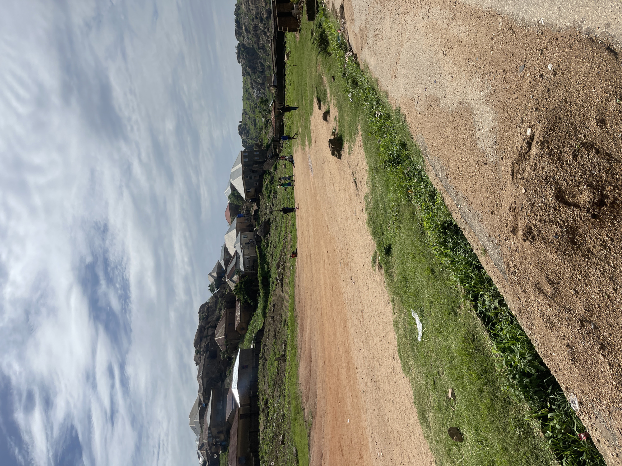 The image depicts a view of a settlement built into a rocky hillside in Jos, Nigeria, under a partly cloudy sky. In the foreground, a dirt path with loose gravel and sand is visible on the right, bordered by a shallow trench filled with green vegetation. To the left of the path is a large expanse of reddish-brown earth, likely a playing field or open area, with patches of green grass along its edges. In the middle ground, several figures, appearing to be children, are scattered across the grassy area, with some engaged in activities on the dirt field. Further back, a collection of houses with corrugated metal roofs are built into and on top of the rocky terrain. The houses are varied in size and construction, with some appearing more modern than others. The landscape is characterized by the prominent rock formations that dominate the hillside.
