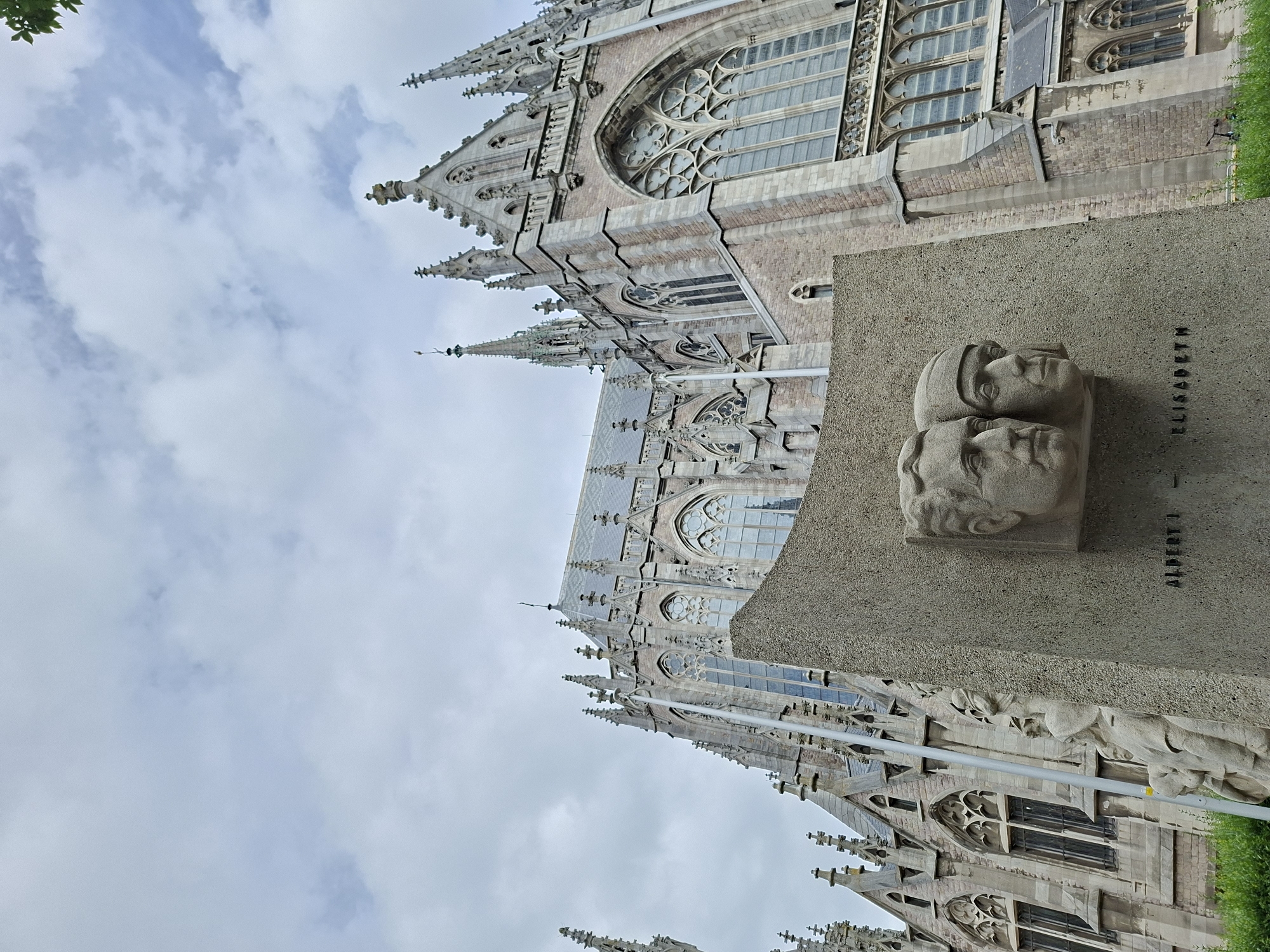The image shows a low-angle view of a monument with two sculpted heads of a man and a woman, along with relief carvings of other figures, in front of the Church of Saint Peter and Saint Paul in Ostend, Belgium. The monument's base is inscribed with "ALBERT I" and "ELISABETH". The church, an example of Neo-Gothic architecture, features spires, pointed arches, and large stained-glass windows. The sky is overcast with visible clouds. Green foliage is present in the lower foreground.