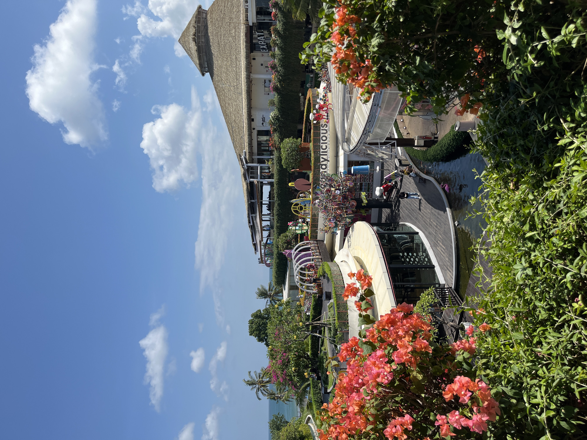 This is a wide-angle, eye-level shot of an outdoor shopping and entertainment complex in Kuta, Indonesia. The sky is bright blue with scattered white clouds. In the foreground, lush green foliage and vibrant orange-pink bougainvillea flowers frame the scene.
The complex features several multi-level buildings with a mix of architectural styles. A large, circular building with a thatched roof is prominent in the background. In front of it, a modern, curved structure with glass walls houses shops. A decorative archway with purple lighting stands in the middle ground, leading to an area with colorful decorations, including what appears to be a tree adorned with candy-like items. A small pond with several koi fish and a pink inflatable flamingo is visible in front of the curved building.
Several people are present: a few individuals are seen walking on the wooden boardwalks and sitting near the pond, while others are visible inside the shops or on balconies. A decorative signage that reads "candylious" is visible. The overall atmosphere suggests a popular tourist destination with a vibrant and colorful design, blending natural elements with modern architecture.