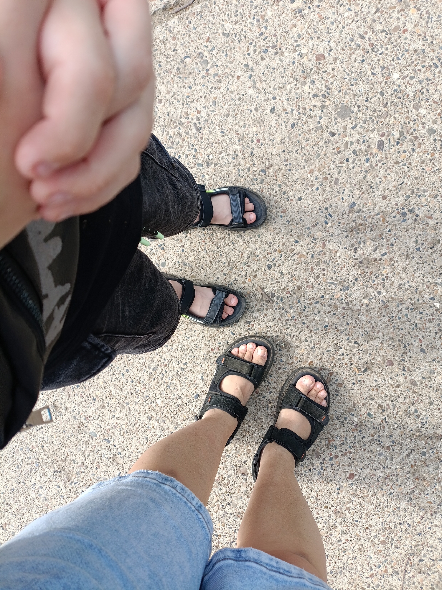 The image shows a ground-level view of three pairs of feet wearing sandals on a concrete surface. The upper pair of feet wears black sandals with gray accents and are covered by black denim jeans. The middle pair of feet wears black sandals. The bottom pair of feet wears black velcro sandals and are covered by light blue denim shorts. The concrete surface is light gray with embedded small pebbles. The upper left corner of the image shows a partial view of a hand in a fist, and what looks like a black jacket. The location of the image is not discernible from the picture. There is no information to suggest the location is Chaikovsky, Russia.