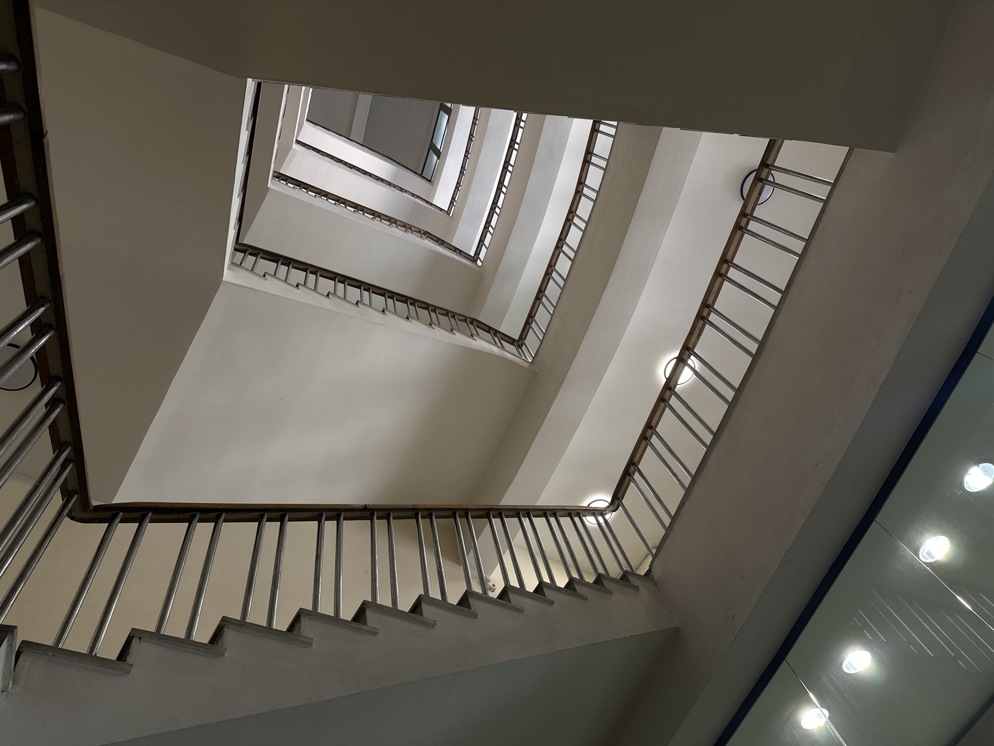 The image shows a view looking straight up inside a stairwell. The stairwell is primarily white with metal and wooden railings. The stairs rise in a square spiral, with windows visible at the top. The location is Yerevan, Armenia. There are no people visible in the image. The environment is indoors, and the lighting is artificial.