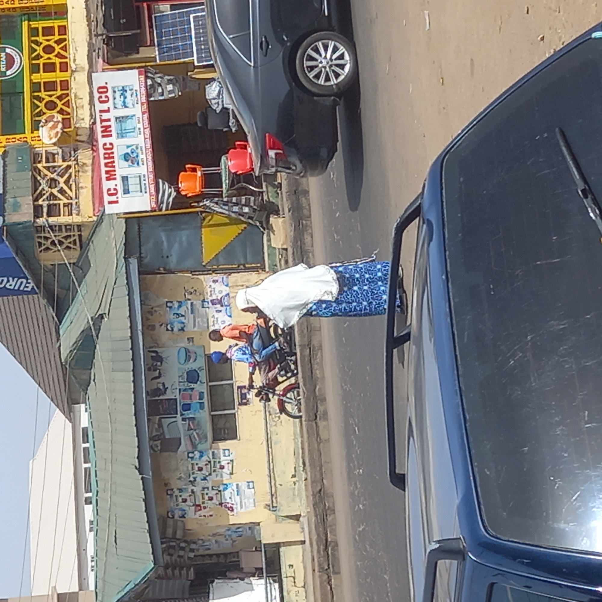 The image presents a daytime street scene in Dadi Riba, Nigeria, viewed from a slightly elevated perspective, possibly from a vehicle. The primary focus is on the street and buildings lining one side, with a blue vehicle partially visible in the immediate foreground.
On the paved street, a black car with silver, spoked wheels is visible, either parked or slowly moving. Clear shadows indicate bright daylight and good weather conditions.
Several individuals are present. In the mid-ground, a person wearing a white head covering and a blue patterned skirt or dress over a white top is walking along the street or sidewalk. Further back, another individual in an orange shirt is partially visible near a motorbike, and another person is situated near a red and white motorbike, possibly preparing to ride or disembark.
The buildings lining the street appear to be commercial establishments. One prominent building features a large white sign that reads "I.C. MARC INT'L CO." Below the main name, smaller text and images suggest the sale of appliances like air conditioners, refrigerators, and washing machines. A phone number, "TEL: 08035760247," is also visible on the sign. Nearby, solar panels are displayed, indicating another potential product offering. Another building has a distinctive yellow and green patterned facade, with the word "RITAN" partially visible on it. A blue sign with "EURO" also appears. Orange buckets or containers are stacked outside one of the shops.
The scene captures typical daily activity in a commercial area, with people moving about and shops open for business. No specific event or strong emotions are discernible; the atmosphere appears routine.