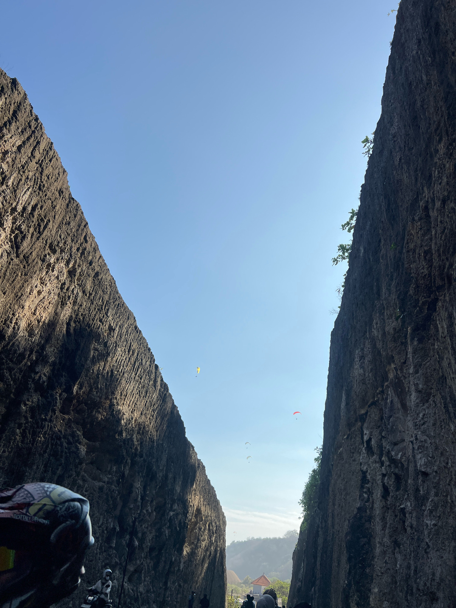 The image depicts a dramatic natural landscape with towering, rough-textured rock formations on either side, framing a clear blue sky. Several paragliders, small colorful shapes against the sky, are visible, indicating an outdoor recreational activity. In the foreground, partially obscuring the view, is the helmet and shoulder of a person, suggesting the image was taken from the perspective of someone on a motorcycle or similar vehicle. Further in the background, a few figures are seen near a small building, and a hazy, tree-covered hillside hints at the wider environment. The scene is likely located in Kuta Selatan, Indonesia, known for its stunning cliffs and adventure activities.