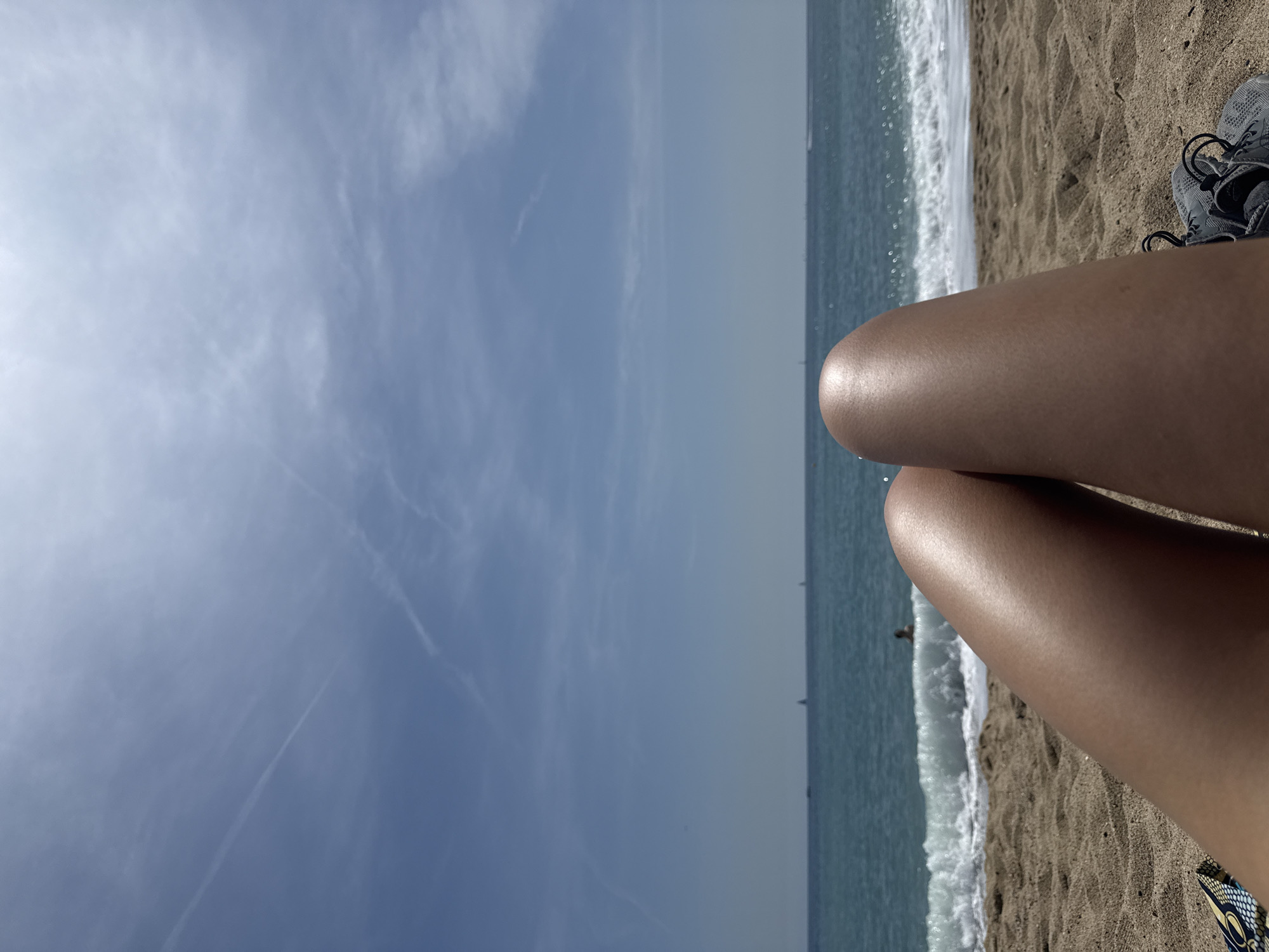 The image depicts a sunny day at a beach. A person is reclining on the sand, with their legs and part of their shoes visible in the foreground. The background shows the ocean with waves crashing on the shore. The sky is mostly clear with some wispy clouds. Given the context of Barcelona, Spain, it is likely that this photo was taken at one of the city's beaches, such as Barceloneta or Nova Icaria.
