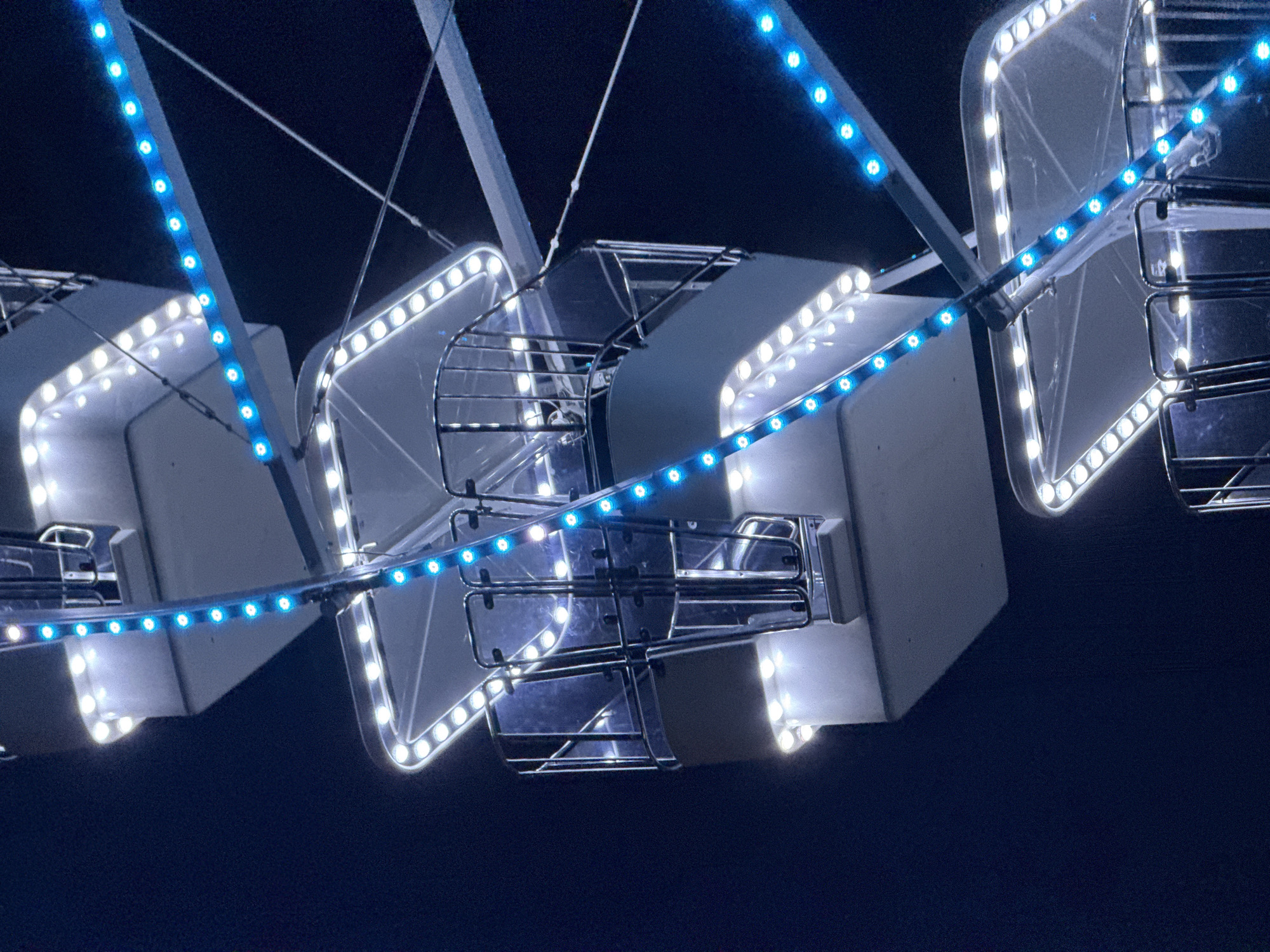 The image shows the illuminated gondolas of a Ferris wheel at night. The gondolas are white with metal railings and clear glass panels. They are decorated with strings of bright white and blue LED lights. The Ferris wheel structure itself is visible, with supporting beams and cables against a dark night sky. The scene appears to be part of an amusement park or festival, likely in the city of Budva, Montenegro, given the context. No people are visible in the image, and the focus is on the illuminated ride and its structure.