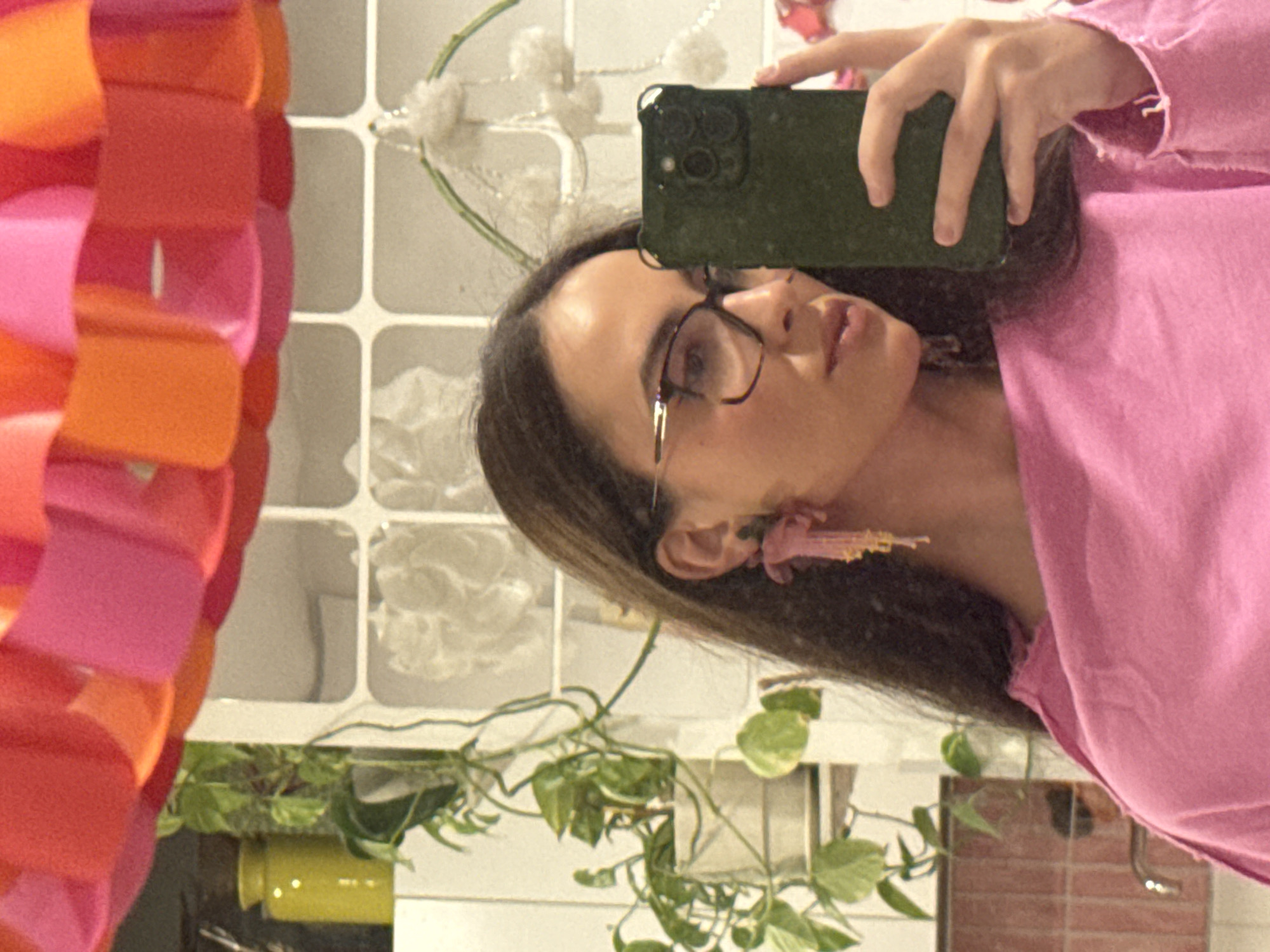The image is a sideways mirror selfie taken by a person, likely female, in an indoor setting. The individual is positioned vertically, with their head to the right and body extending downwards. They have long dark hair, wear glasses, and are dressed in a pink long-sleeved top. A distinctive pink and gold floral-like dangle earring adorns their visible ear. The person holds a dark green smartphone, equipped with a multi-lens camera system, in their right hand, pointed towards the mirror to capture the reflection.
The background, visible in the mirror's reflection, features a white tiled wall with a grid pattern. Below this, green trailing plants, consistent with species like pothos, are visible, suggesting indoor greenery. A yellow pot or vase is partially seen behind the plants. To the far left of the reflection, a vibrant, segmented object in shades of pink, orange, and red stands out, possibly a decorative lampshade or a sculptural piece. Other faint reflections include white, fluffy decorative elements on a wire frame in the upper background.
The lighting is bright and even, indicating an indoor daytime environment. The person maintains a neutral expression, focused on the act of taking the photograph. There is no visible text present in the image. This scene is located in Krasnoyarsk, Russia.