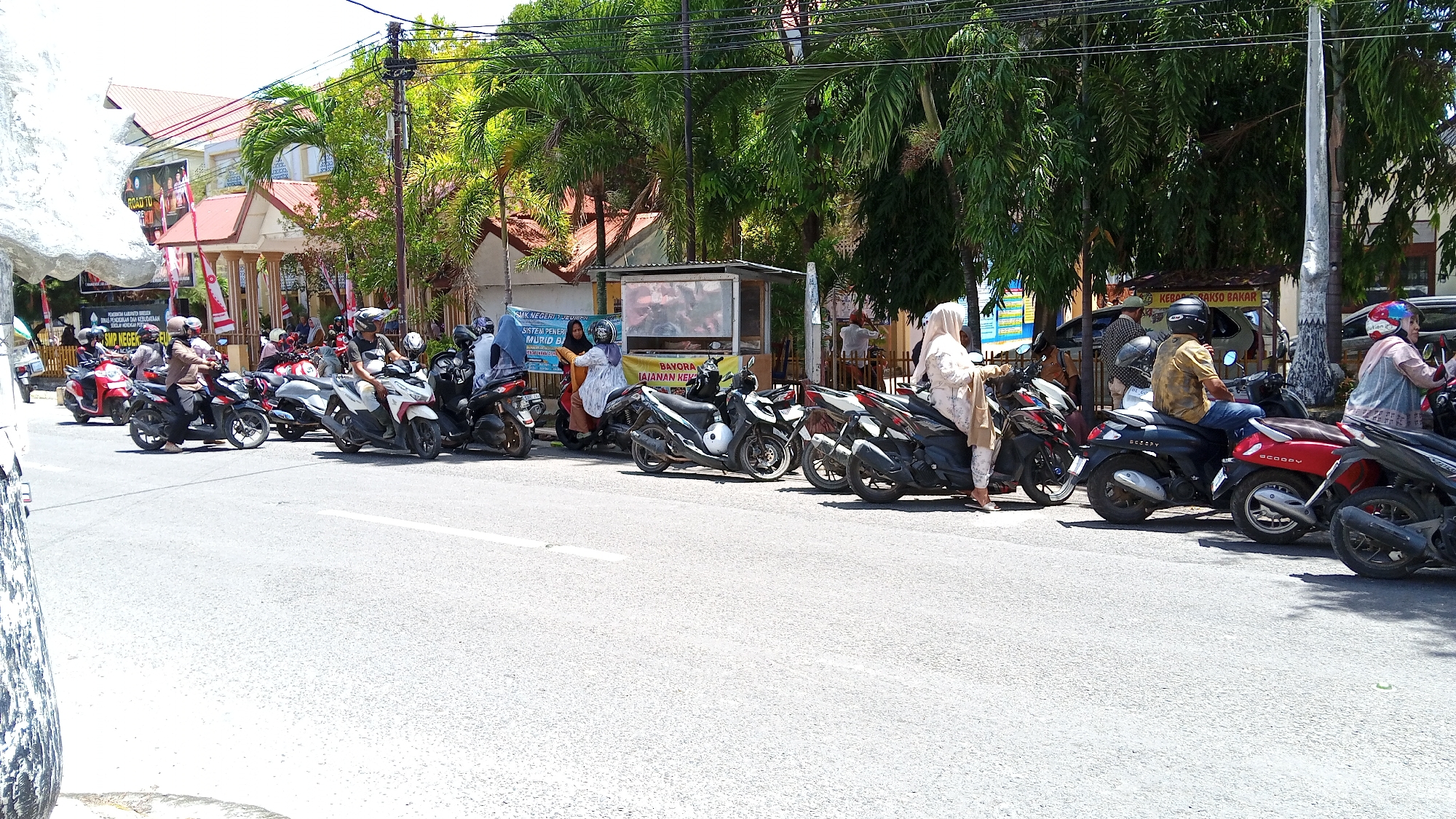 The image depicts a street scene in Pulokiton, Indonesia, with a row of parked motorcycles lining the side of the road. In the background, there is a building with a red roof, possibly a school or government building, adorned with banners and flags. Several people are seen on or near the motorcycles, some wearing helmets and headscarves. The atmosphere appears to be that of a busy street, with the presence of palm trees and a utility pole indicating a tropical or sub-tropical urban environment. The motorcycles are of various makes and models, and some appear to be of the scooter type. The road surface is asphalt, and there are white road markings visible. There is also a small food stall visible in the middle background, suggesting commercial activity. The lighting indicates a bright, sunny day.