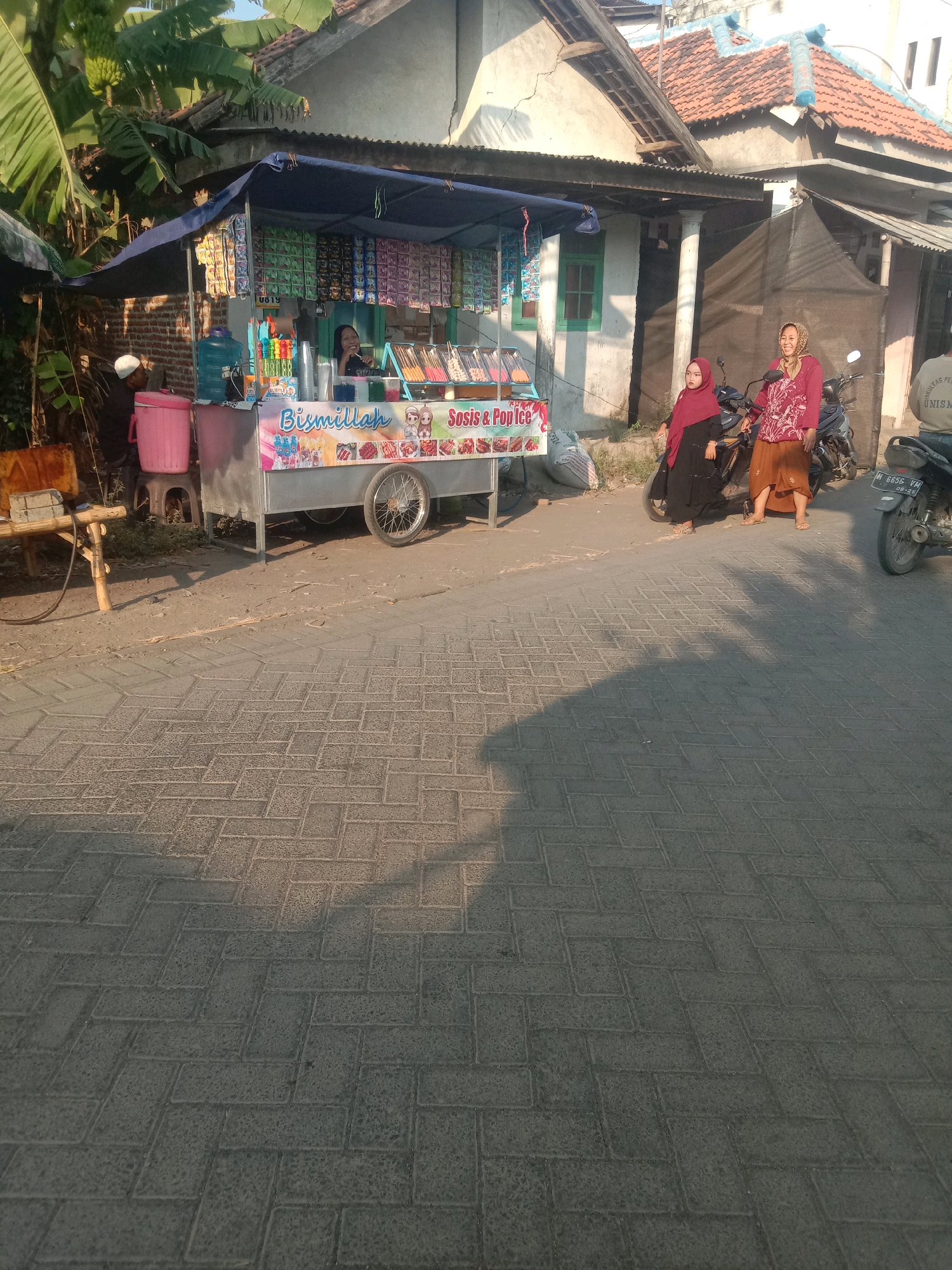 A street-level view captures a food cart in Selotambak, Indonesia, bathed in late afternoon sun. The cart is adorned with colorful banners, displaying items like "Bismillah," "Sosis & Pop Ice." Behind the cart, a woman in a headscarf smiles as she attends to a customer. To the right, two women in hijabs stand near a parked motorcycle. A man in a white cap sits near a large pink cooler, while a bamboo bench and table are visible to the left. The background shows a house with a tiled roof and lush banana trees. The street is paved with interlocking bricks, reflecting long shadows from the setting sun.