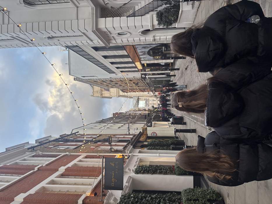 An upward-tilted street view features two girls in black puffer jackets. The street is lined with brick and white buildings, some adorned with greenery. String lights hang across the street, converging in the distance where a crowd of people is visible. A sign reads "agnès b. PARIS." The sky is cloudy with patches of blue.