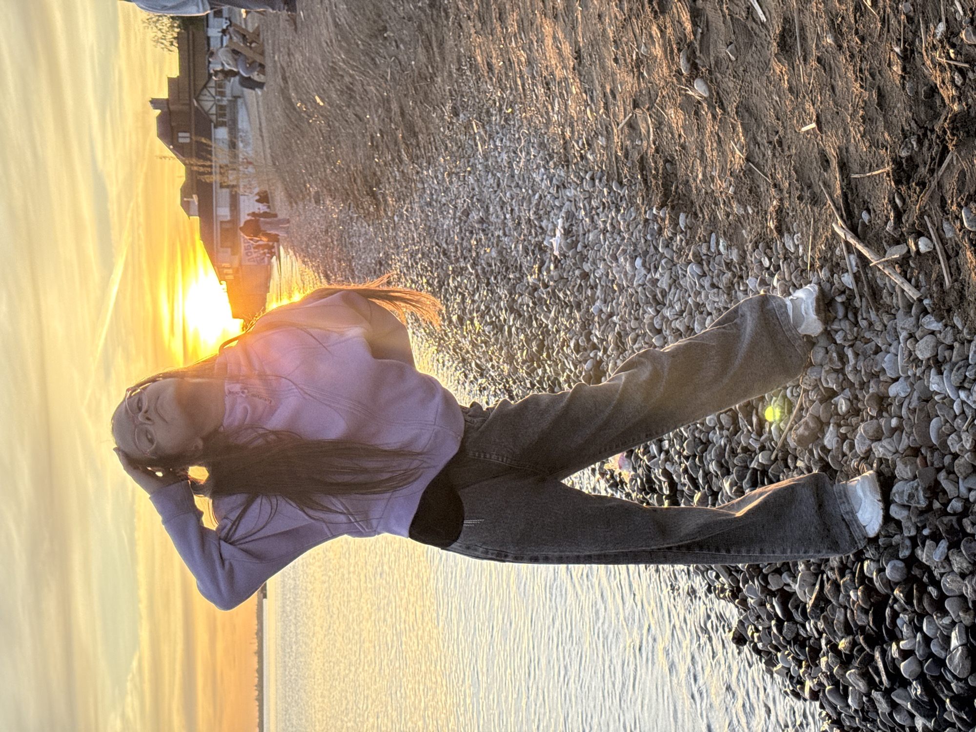 A person stands on a rocky beach, possibly in Unknown, Unknown, with a body of water to their left and a land mass behind them. The individual is wearing a light purple hooded jacket, dark gray jeans, and white sneakers. Their posture is somewhat posed, with one hand on their head. The background includes a sunset or sunrise illuminating the scene, a building, and other individuals further down the beach.