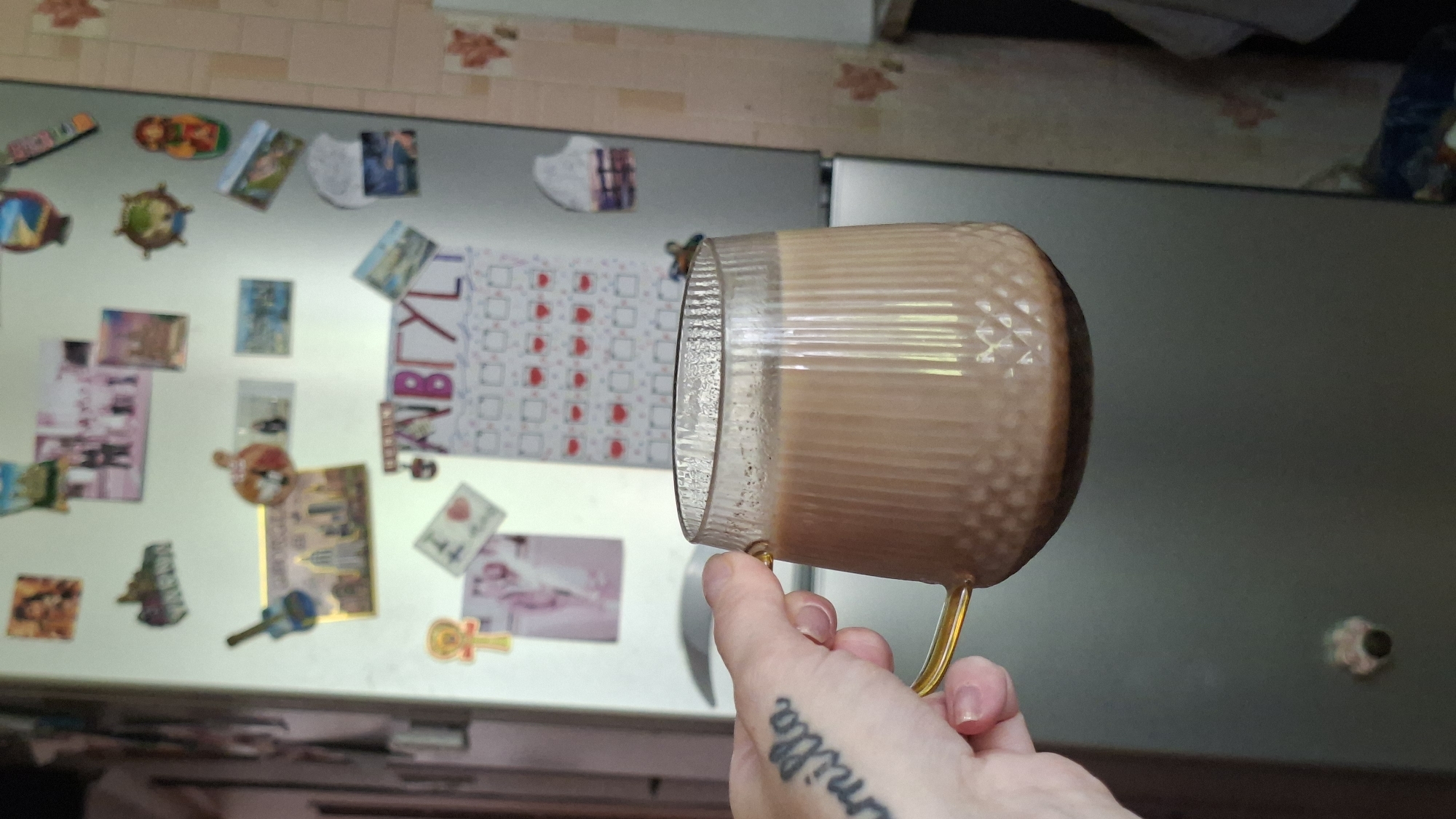 A person's hand, tattooed with the word "amilla," holds a ribbed glass mug filled with a light brown beverage, possibly coffee or tea, against the backdrop of a refrigerator covered in magnets and a calendar. The refrigerator is silver and appears to be in a kitchen setting. The magnets on the refrigerator depict various images, including photographs, landmarks, and decorative items. A calendar with the letters "АВГУСТ" (August in Russian) is also visible, along with small red heart shapes. The surrounding environment suggests a domestic space, likely a kitchen. The image does not provide any information that would place it in the city of Myadzyel, Belarus.