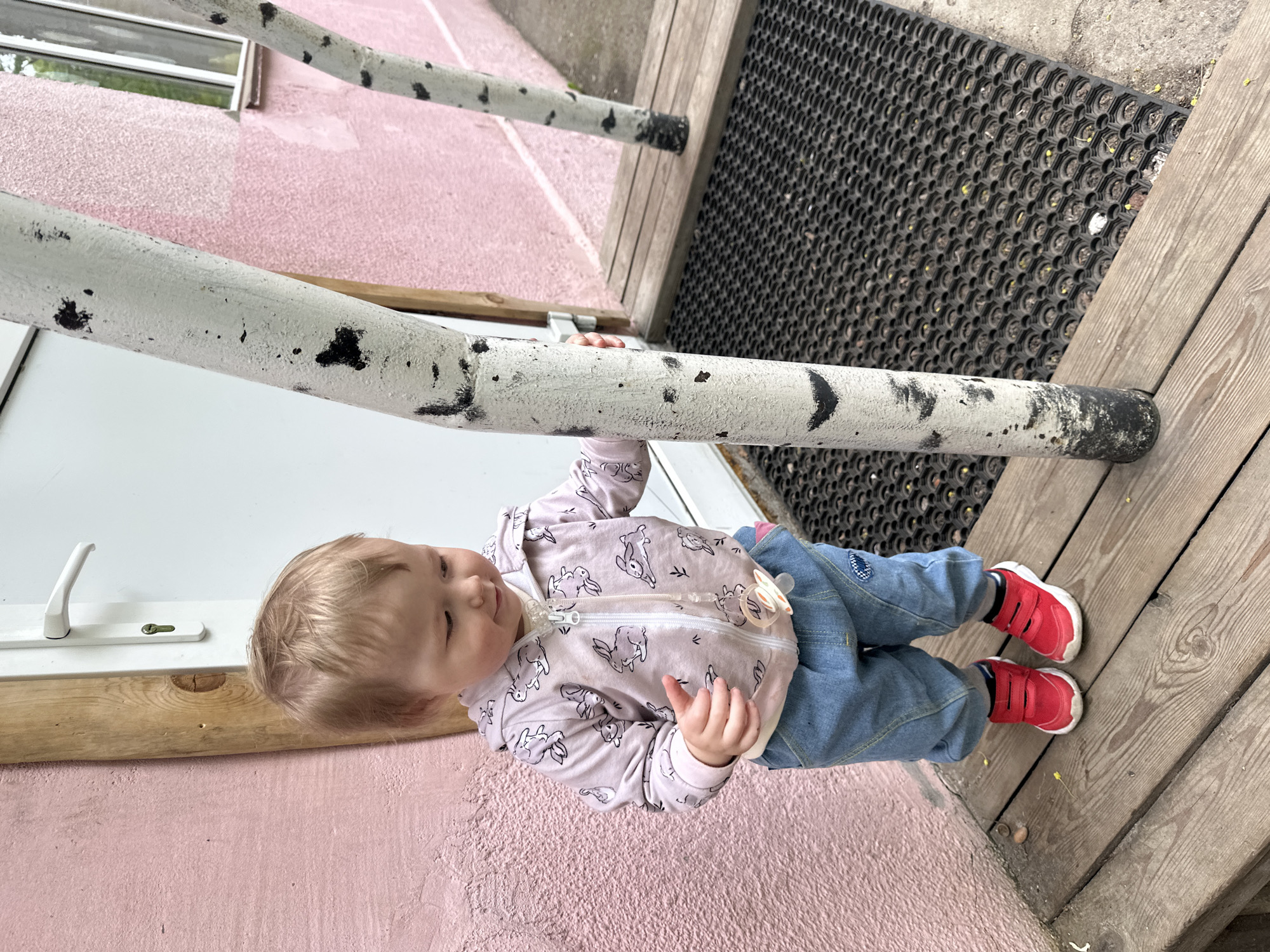 A toddler stands near a pink wall next to a wooden porch with a black rubber mat. The child wears a bunny-patterned jacket, blue jeans, and red shoes. They are grasping a white pole with black markings that runs horizontally across the image. The scene appears to be outdoors, potentially in a residential area. No details indicate the location is Nizhny Novgorod, Russia.