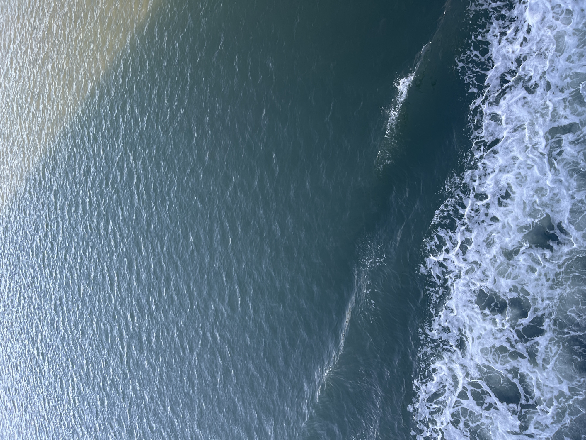 This image depicts a dynamic body of water, likely a river or estuary, with two distinct water types meeting. The lower left of the frame is dominated by churning white foam and waves, indicative of a moving vessel's wake. The water transitions to a deep blue-green hue. The upper right portion of the image shows a contrasting lighter, murkier water, suggesting a confluence with another water source, possibly carrying sediment or having a different temperature. The surface of the water exhibits a gentle ripple pattern throughout, catching ambient light. There are no visible people, structures, or specific landmarks that would allow for the identification of the location within Batas Desa Skotong Timur, Indonesia. The scene is an abstract representation of water dynamics.