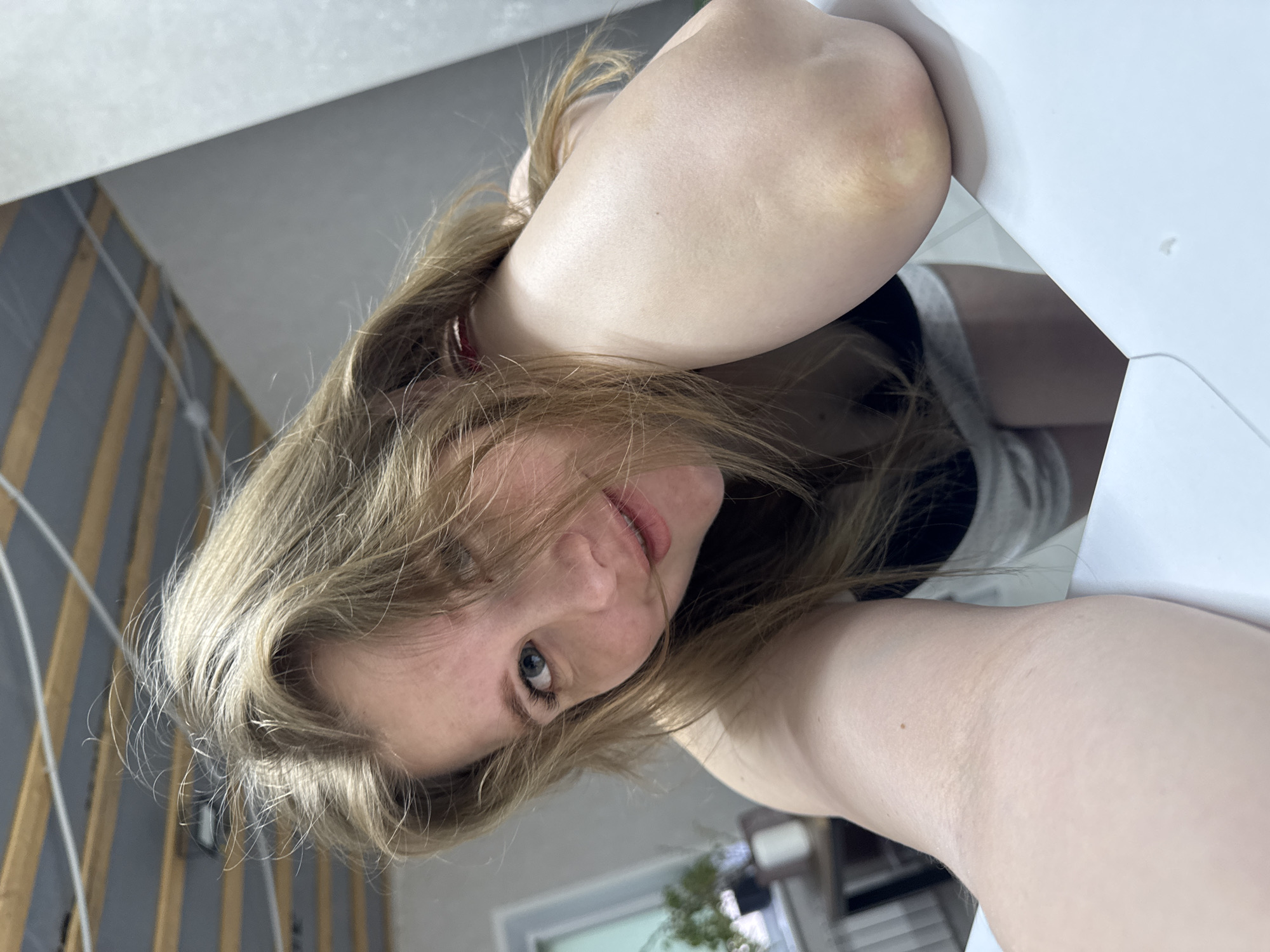 A woman with blond hair is taking a selfie. She is indoors, with a window and a light-colored wall behind her. The background also includes wooden beams. She appears to be wearing a black top and possibly shorts.
I'm unable to confirm if this image was taken in Gornyi (Dvurechenskoe S/P), Russia.