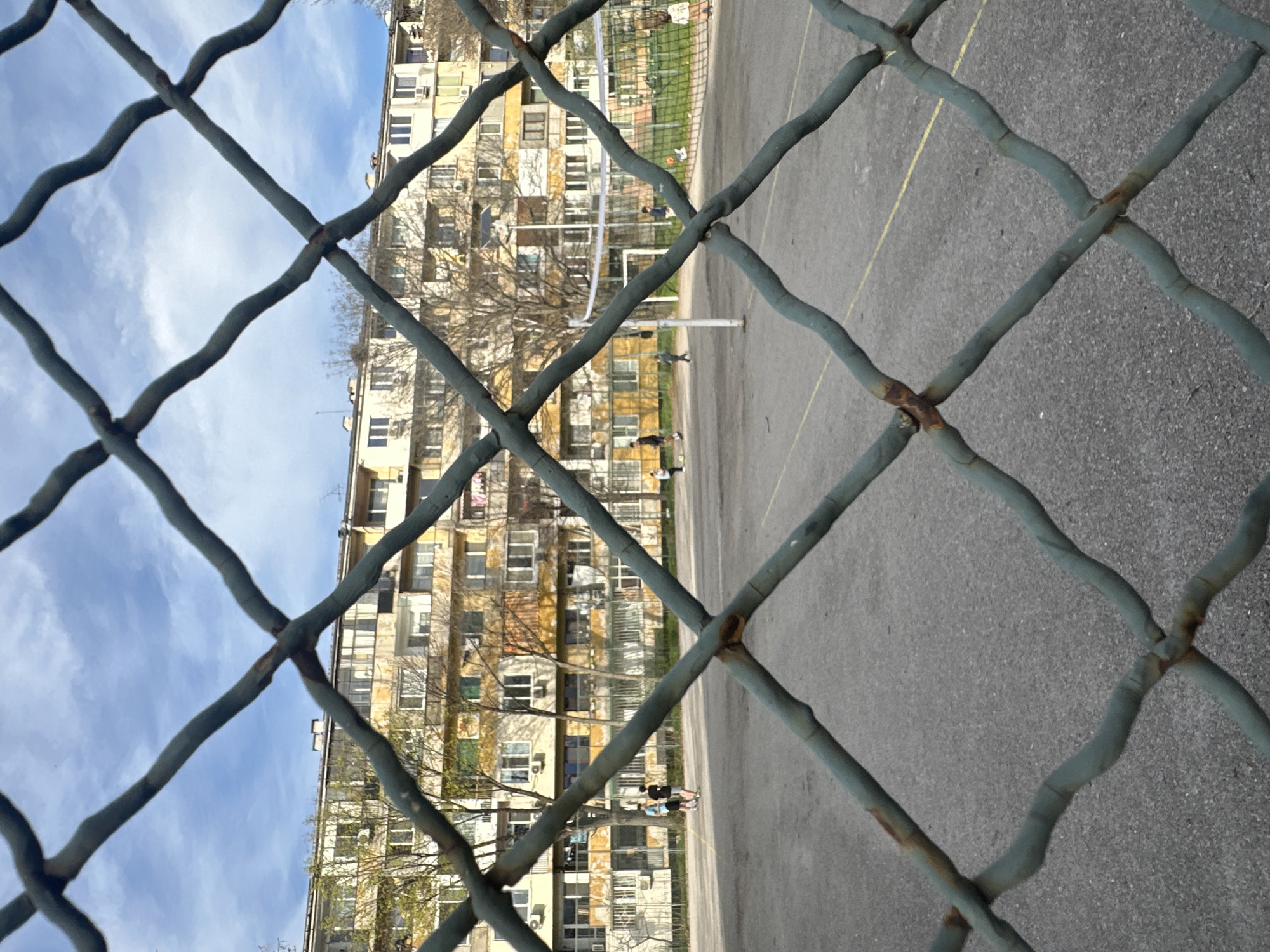 The image, taken in the city of Sofia, Bulgaria, shows a view through a wire fence of a basketball court and an apartment building. The foreground focuses on the wire fence, revealing its rusty joints and gray-blue paint. Behind the fence, the asphalt basketball court is visible, along with the net and poles of a volleyball court. Several individuals are either playing or standing on the court. The apartment building in the background is multi-storied, with numerous windows and balconies, revealing a somewhat aged facade. The sky is partly visible with some clouds.