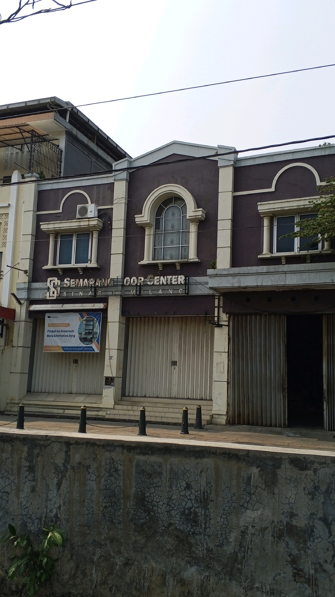 The image shows the exterior of a building that houses "SEMARANG FLOOR CENTER" and "SINAR SEMARANG". The building is a two-story structure with a dark purple facade and cream-colored accents around the windows and entrances. The ground floor has two large, white, roll-up doors, indicating that the business is likely a retail or showroom space. Above these doors are windows, one of which is arched and features decorative glass panes. An air conditioning unit is visible on the second story, mounted above a rectangular window. A banner with text in Indonesian is hanging on the left side of the building, partially obscuring the roll-up door and what appears to be a showroom display. The building is situated in an urban environment, with a concrete wall and some vegetation in the foreground, suggesting it might be located near a canal or waterway. The sky is bright and overcast. The scene appears to be in Semarang, Indonesia, based on the name of the business.
