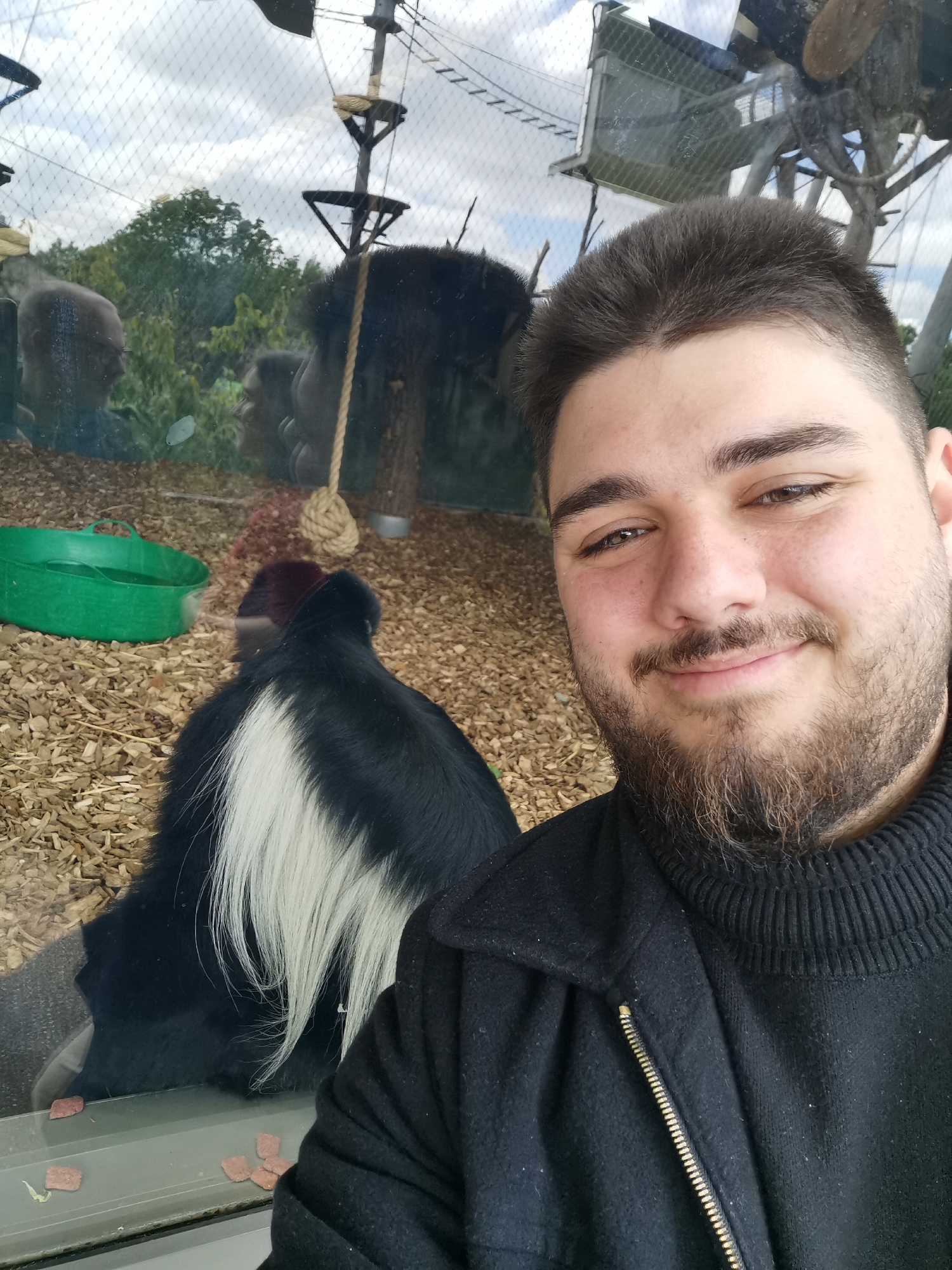 A selfie shows a man with brown hair and a beard. He is smiling at the camera and is wearing a black turtleneck sweater and a black jacket with a gold zipper. In the background, behind a pane of glass, are several black and white colobus monkeys. They appear to be in a zoo habitat, with wood chip ground cover. A green bowl sits to the left, and there is a climbing structure in the distance. The location is unknown.