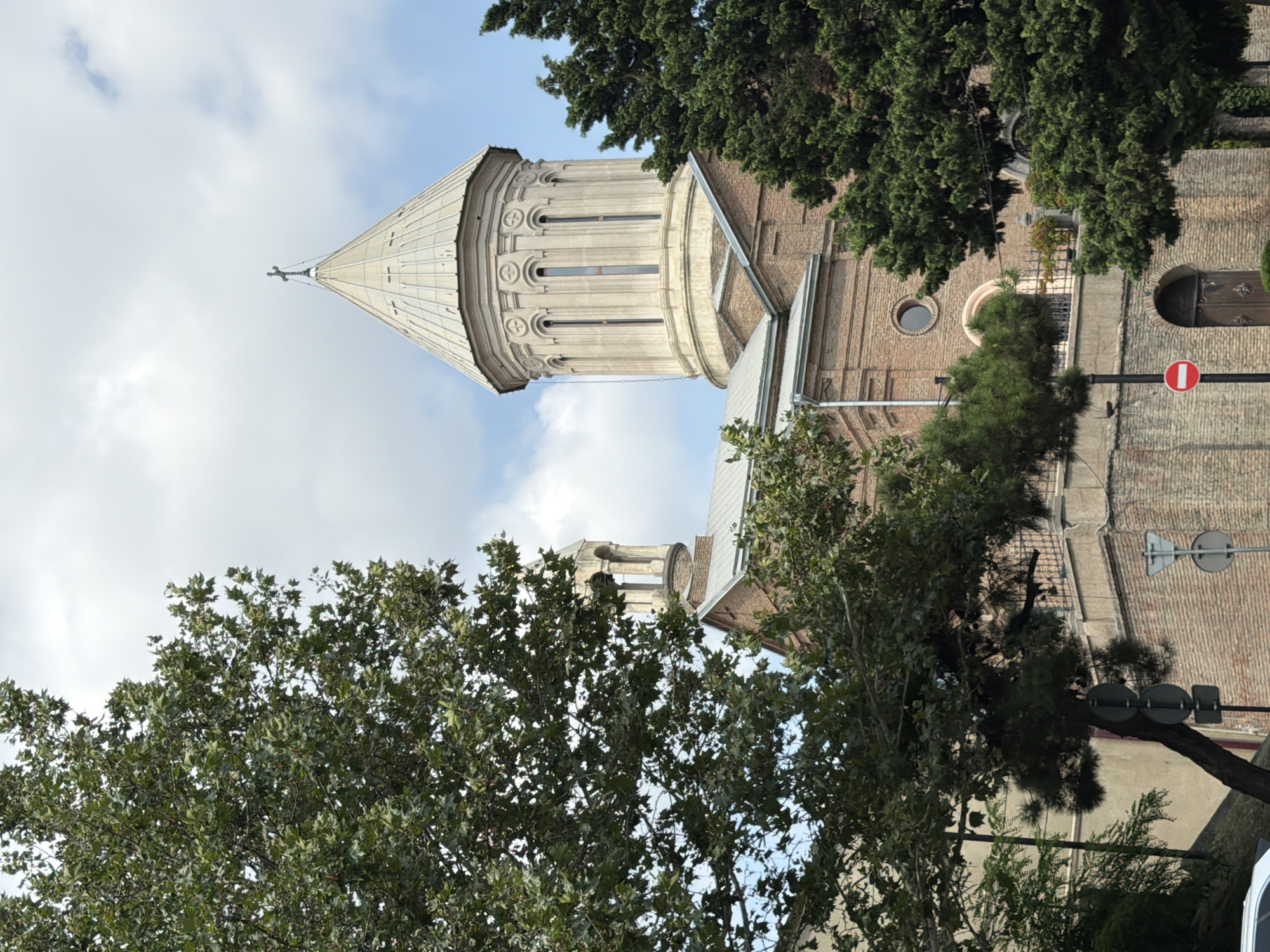 The image shows the Sioni Cathedral of the Dormition in Tbilisi, Georgia. A large, leafy tree with dark green foliage dominates the left side of the frame, partially obscuring the cathedral's bell tower. The cathedral itself is constructed of red brick and features a large, domed roof topped with a cross. The dome is adorned with architectural details and windows. Pine trees are visible on the right side of the image. In the foreground, there are traffic signs, including a "no entry" sign, and a dark wooden door. The sky is partly cloudy.