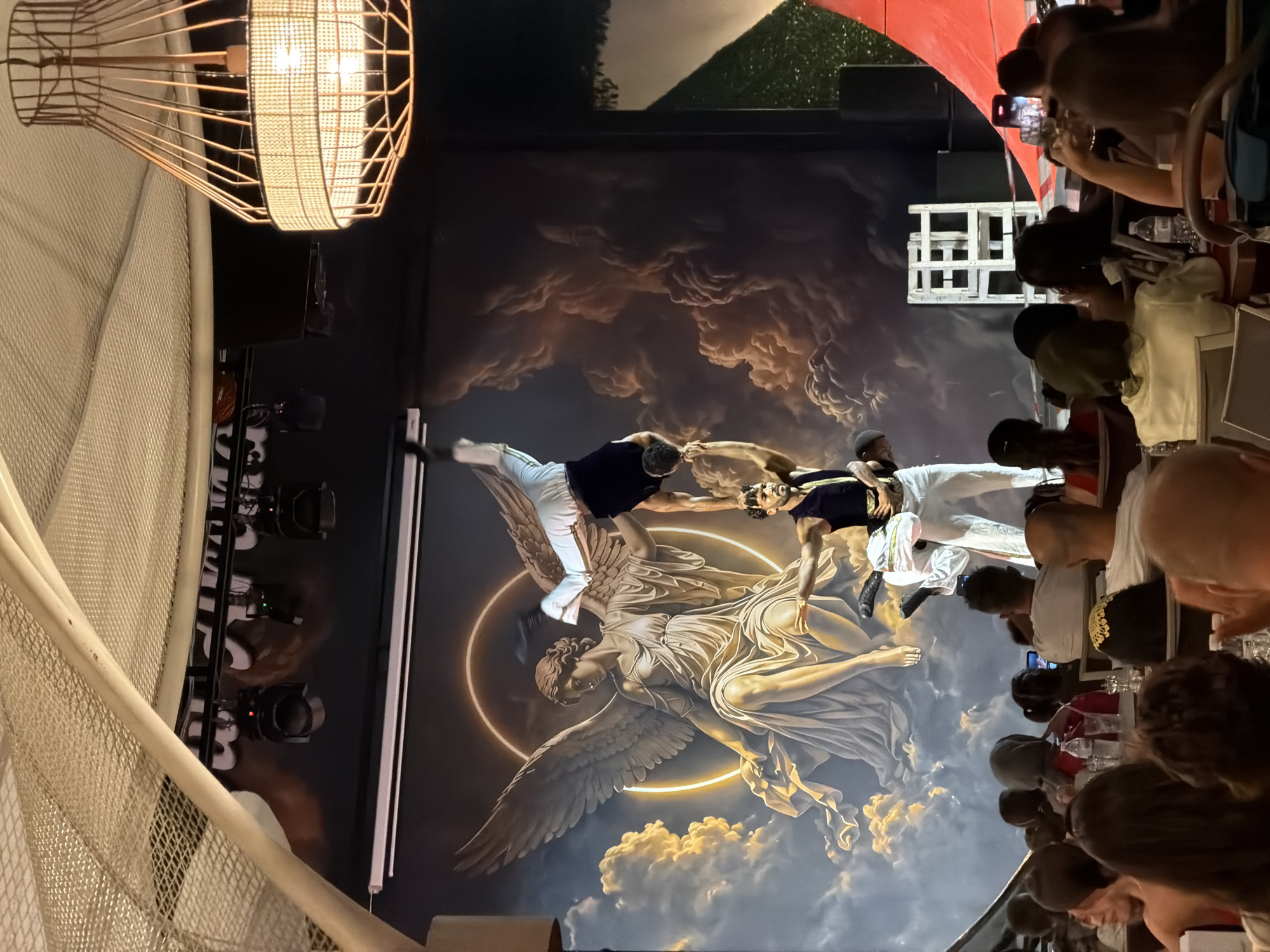 In Manavgat, Turkey, a performance is underway on a stage featuring a mural of an angel with wings spread against a cloudy sky and a golden halo. Three acrobats are performing a human pyramid. The bottom acrobat is standing, wearing a dark vest and white pants. The middle acrobat is in a handstand on the bottom acrobat's shoulders, wearing a white shirt and dark pants. The top acrobat is in a handstand on the middle acrobat's shoulders, wearing a white outfit. Spectators are seated at tables in the foreground, some taking photos with their phones. A large, woven lampshade hangs from the ceiling.