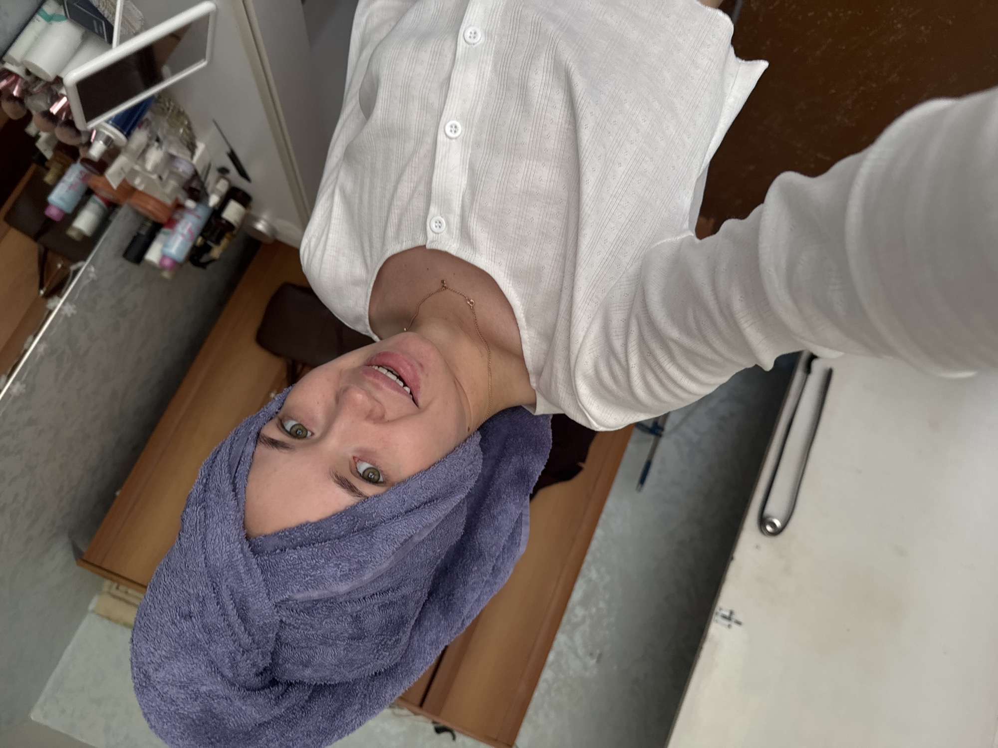 A young female is depicted in a selfie, smiling directly into the camera from an indoor residential setting in Chaikovsky, Russia. Her head is wrapped in a lavender terrycloth towel, turban-style. She wears a white, long-sleeved, round-neck top with a subtle textured pattern resembling tiny dots within vertical ribbing, featuring a five-button placket down the front. A thin gold-colored necklace with small charms adorns her neck. Her eyes are green, and she has light skin with minor blemishes visible around her mouth.
The background elements, from left to right, include a white door or wardrobe with a chrome handle, a tall brown wooden wardrobe or cabinet, and a wall covered in textured, light gray wallpaper. To the right, a white vanity table is visible, cluttered with numerous cosmetic and personal care items, including various bottles, jars, makeup brushes in holders, a small white standing mirror, and a tablet device. A portion of a mirror reflecting the subject's shoulder and parts of the room is visible on the far right.