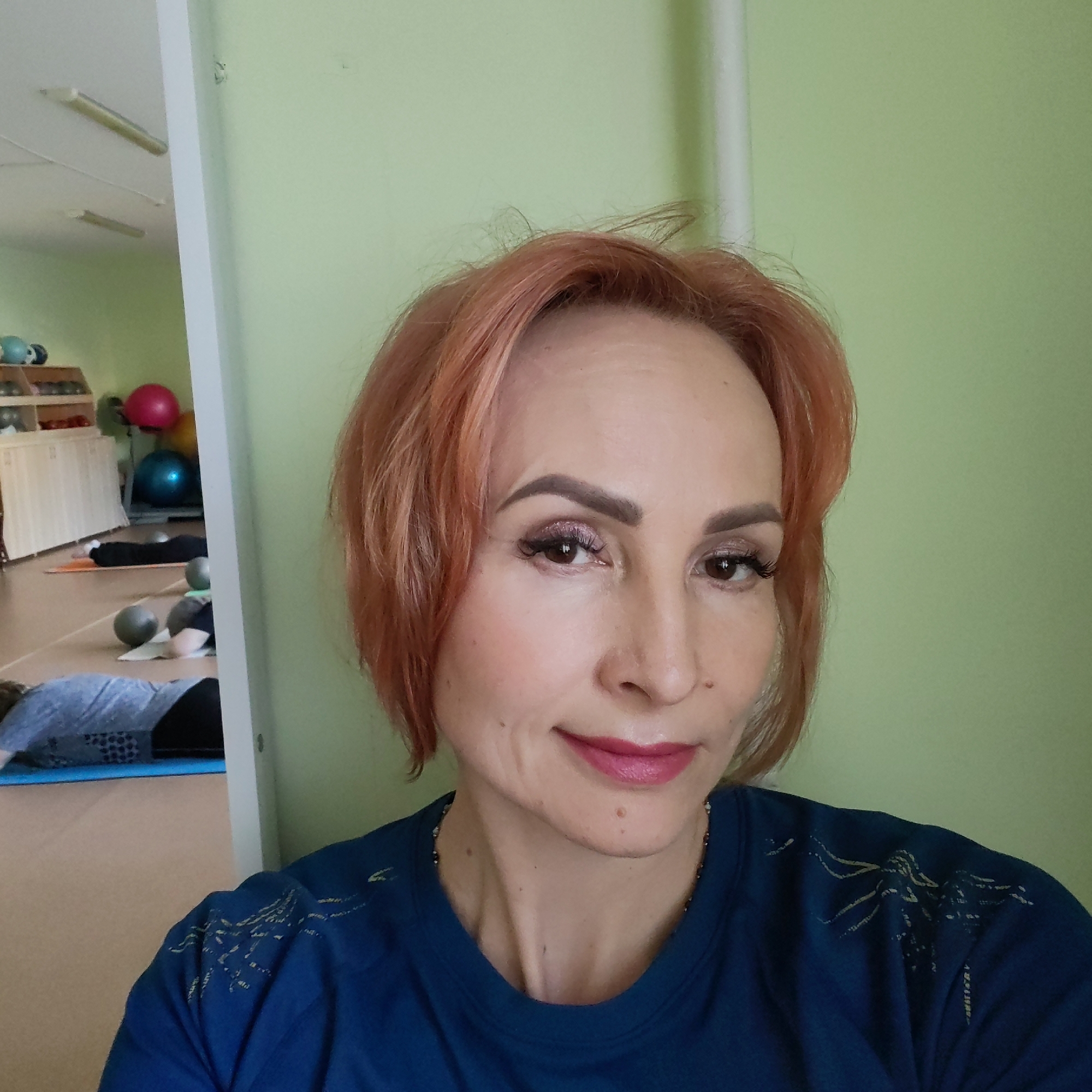 The image is a selfie taken by a middle-aged woman with short, reddish-blonde hair. She has fair skin, visible wrinkles, and wears makeup including eyeshadow, eyeliner, and lipstick. She is wearing a blue t-shirt with a design in light gold. The background is a light green wall, next to a white doorframe. Through the doorway, a room can be seen with exercise balls, mats, and people lying on the floor, suggesting a gym or fitness studio. Based on the location data, the image was likely taken in a fitness establishment in Chaikovsky, Russia. The selfie is posed, with the woman looking directly at the camera and smiling slightly.