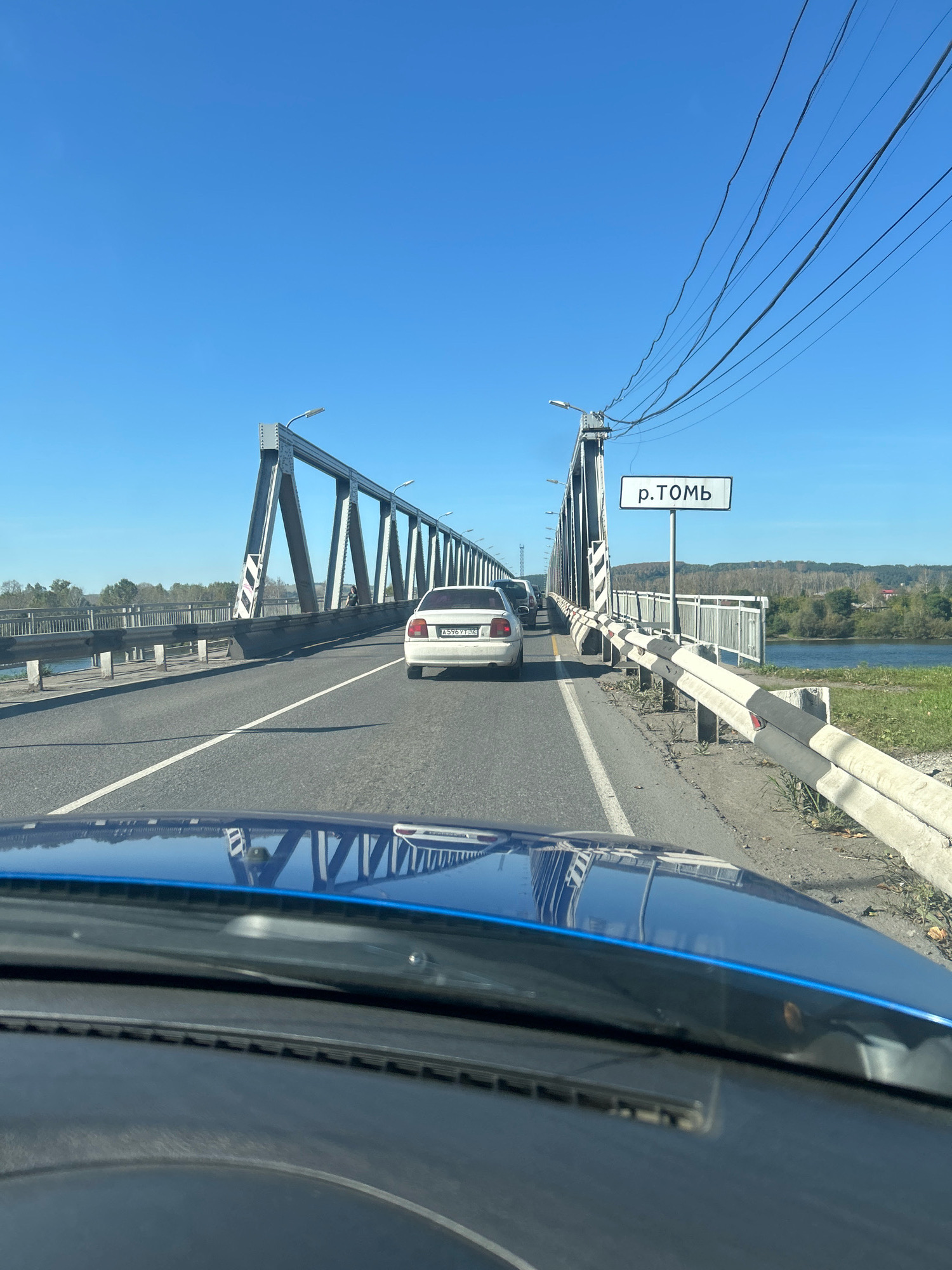 The image shows a view from the interior of a vehicle, looking out over the hood and windshield, towards a bridge spanning a river. The bridge is a steel truss structure with multiple spans and is carrying vehicular traffic. A white sedan is visible in front of the vehicle, traveling on the bridge. There are guardrails on either side of the road, and a sign is posted that reads "р. ТОМЬ," indicating that the bridge crosses the Tom River. The sky is clear and blue, and the surrounding landscape appears to be rural, with trees and vegetation visible on the banks of the river and in the distance. The reflection of the bridge and the sky can be seen on the hood of the vehicle. The context suggests this is a view from the Tom River bridge in Novokuznetsk, Russia.