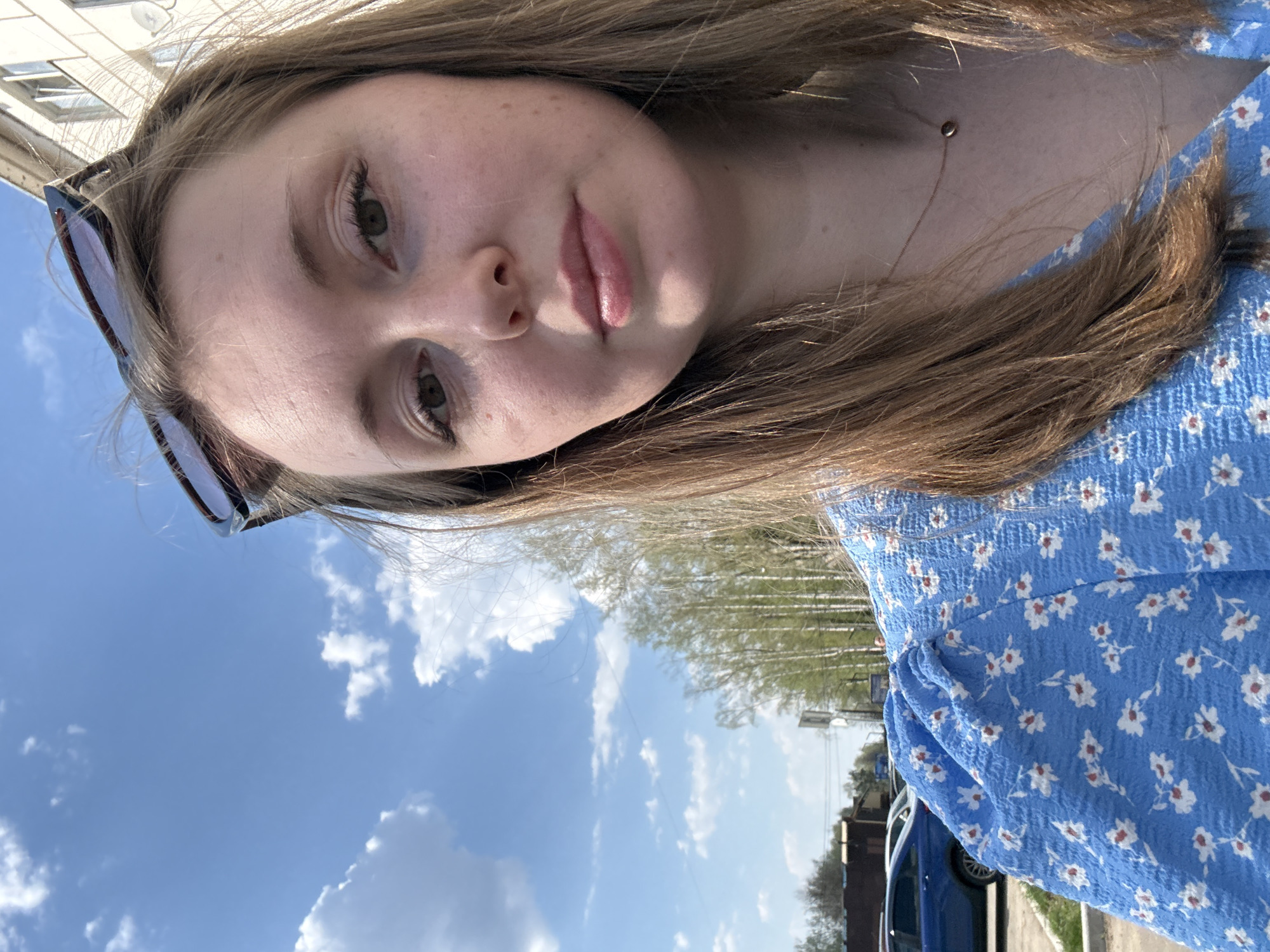 The image depicts a young woman with light skin, brown hair, and a blue floral-patterned dress. She is wearing sunglasses on her head and a delicate necklace. The background shows a clear blue sky with scattered clouds and trees in the distance. A building and a blue car are partially visible. The image appears to be a selfie taken outdoors in an urban environment.
