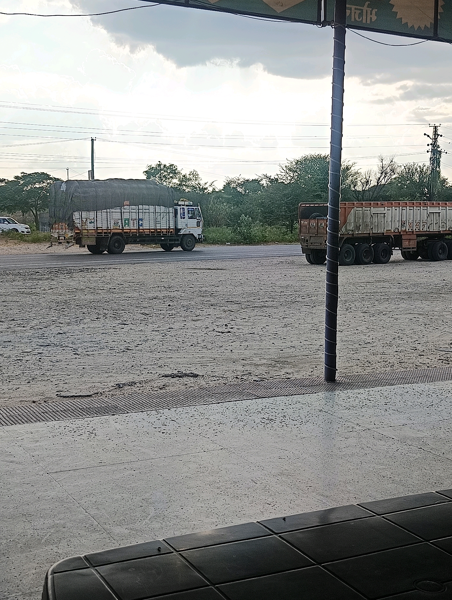 This image captures a roadside scene in Chhapri Bari, India. Two trucks are visible on a road. The truck on the left is white with a large, dark tarp covering its cargo. The truck on the right is a flatbed trailer, its sides are partially obscured by a pole in the foreground. The road is flanked by a sandy, gravelly area and sparse vegetation. Power lines crisscross the sky, and a tall pole with a coiled wire runs vertically through the center of the frame. The sky is overcast with dramatic clouds. The foreground features a textured concrete surface and a dark, tiled object, possibly part of a structure.