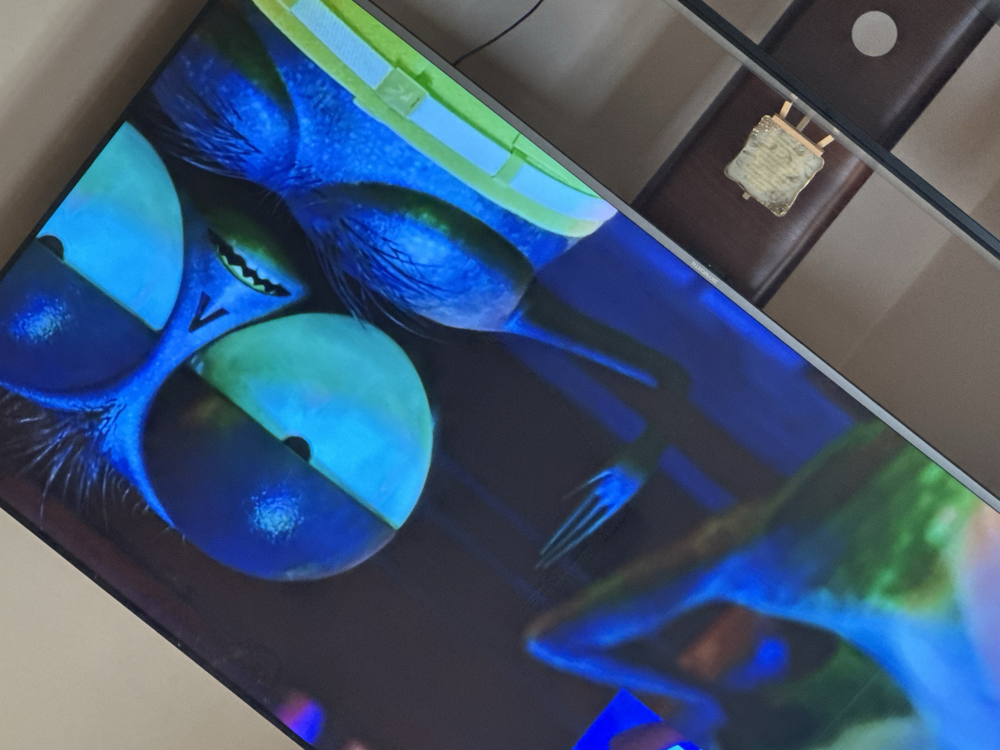 The image shows a close-up of a television screen displaying a brightly colored image of what appears to be a character with large eyes, sharp teeth, and an unnatural blue hue. The TV appears to be placed on a table. The screen also partially reflects another object on the table, possibly an award of some sort, and a dark-colored surface. The location is not discernible from the visual information provided.