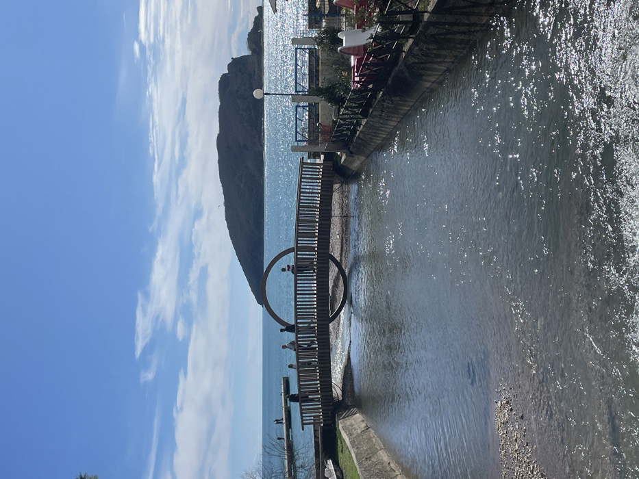 This image captures a coastal scene in Budva, Montenegro, centered on a pedestrian bridge spanning the Grđevica river/canal, approximately 200 meters from its confluence with the Adriatic Sea. The prominent landmass in the background is Sveti Nikola Island.
The foreground features a narrow, concrete-lined waterway flowing from left to right towards the sea. The water's surface reflects sunlight, and its shallow areas reveal a gravel bottom. A wooden pedestrian bridge, approximately 20 meters long, crosses this waterway in the mid-ground. The bridge features vertical wooden railings and a prominent dark, circular metal archway integrated into its structure over the water. Eight individuals are visible walking on the bridge: three on the left side of the arch, two directly beneath it, and three on the right.
To the left of the waterway's mouth, a concrete pier or jetty extends into the sea, where several indistinct figures are standing. Beyond the bridge, the light blue-green sea extends towards the horizon. Sveti Nikola Island, a large, dark, tree-covered landmass, occupies the central background.
On the immediate right side of the waterway, a concrete retaining wall supports a recreational area. This area is enclosed by a dark metal fence and contains multiple red tables, white chairs (some designed as swan-shaped pedal boat seats), and a single white lamp post with a spherical globe light. Green foliage is visible behind the tables.
The sky is a bright, clear blue with scattered white cumulus clouds, particularly in the upper left quadrant. The scene is illuminated by strong sunlight, indicating a clear weather day.
