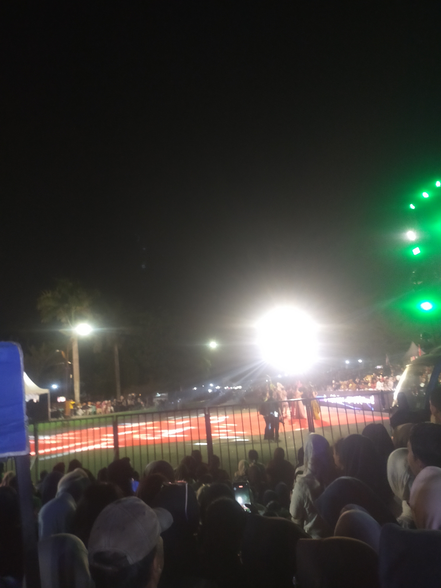 The image depicts a nighttime outdoor event, possibly a concert or festival, in the city of Slawi, Indonesia. A crowd of people is visible, gathered behind a fence. In the background, bright lights illuminate the scene, suggesting a stage or performance area. Some individuals are holding up their phones, possibly to record or take pictures. The atmosphere appears to be festive and lively.