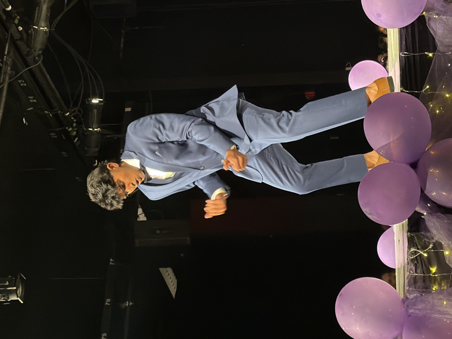 A male individual stands centered on a decorated theatrical stage in Palos Verdes Estates, United States, captured in a dynamic, mid-action pose. He is wearing a light blue three-piece suit, a white dress shirt, a dark bow tie, and tan dress shoes. His dark, wavy hair is styled upwards, and he wears a wristwatch on his left wrist. Both hands are clenched into fists and held near his chest, while his mouth is open. The immediate foreground of the stage is adorned with numerous matte purple balloons, purple mesh fabric, and small string lights. The background is dark, revealing overhead stage lighting fixtures, cables, and numbered structural beams (e.g., '79', '78'). Faint outlines of spectators are visible in the lower right background.